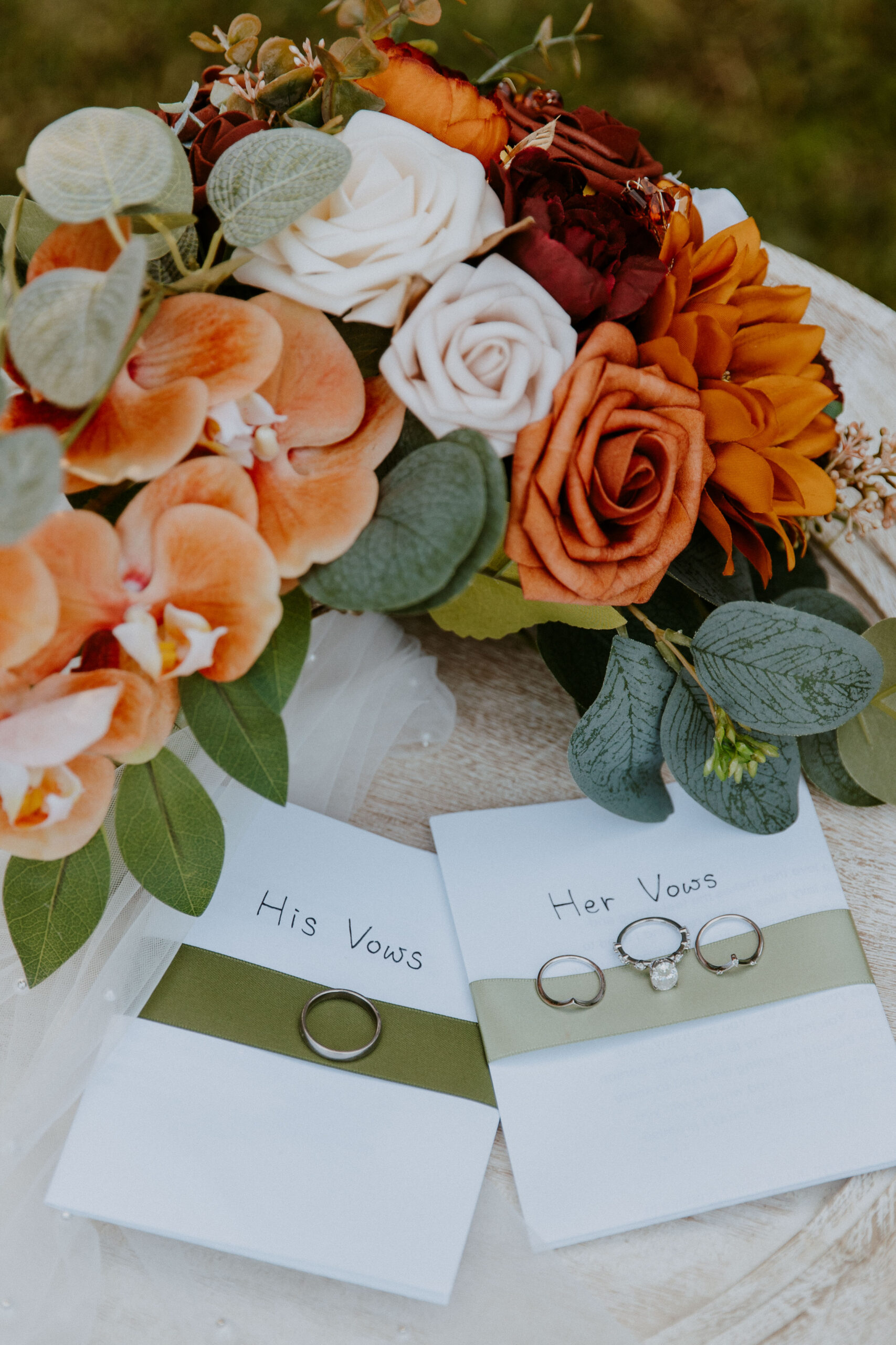A wedding setup featuring vow booklets labeled "his vows" and "her vows" with rings on top, placed beside a bouquet of flowers.