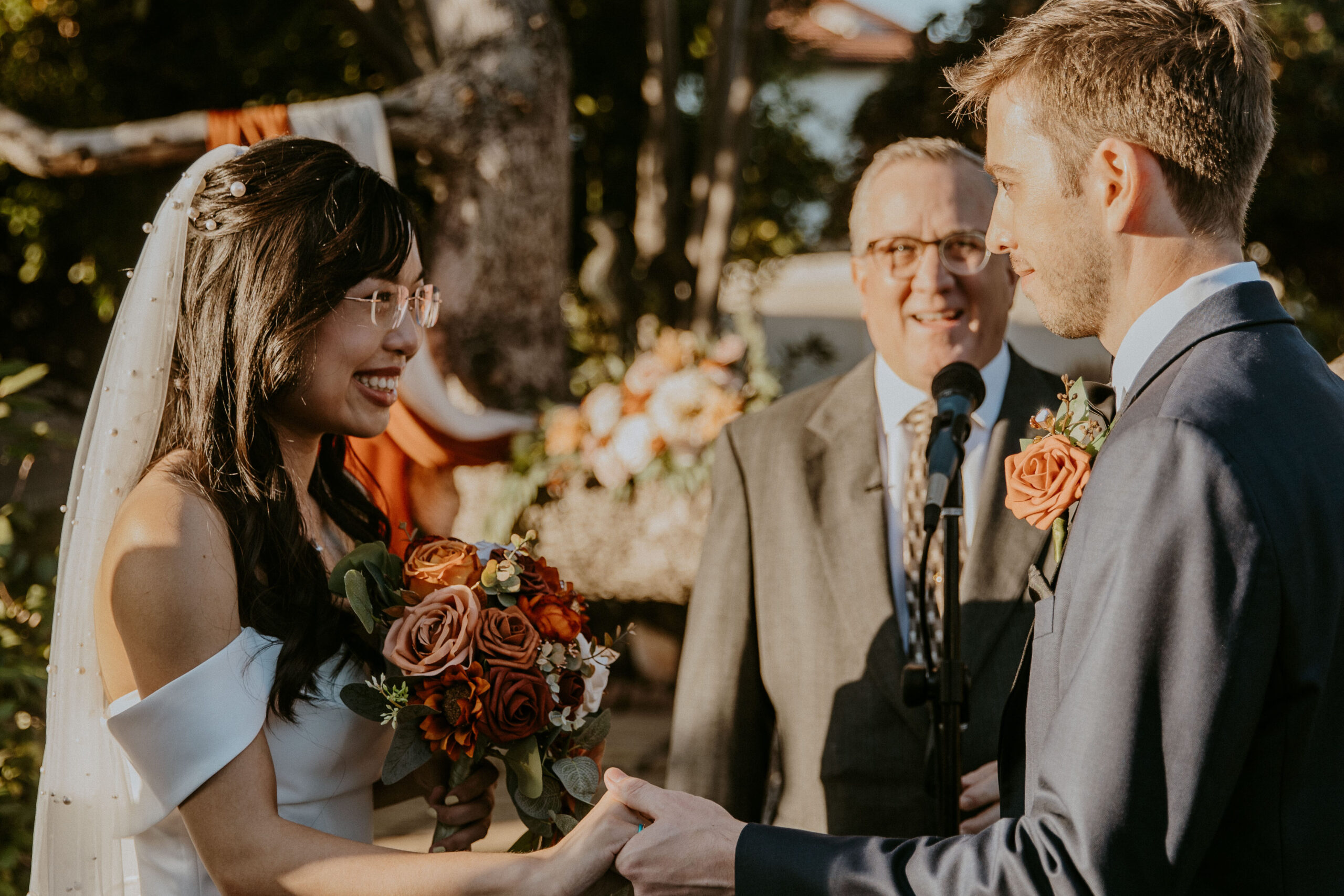 A bride and groom exchange vows at an outdoor wedding ceremony, with an officiant standing in the background at their intimate backyard wedding.