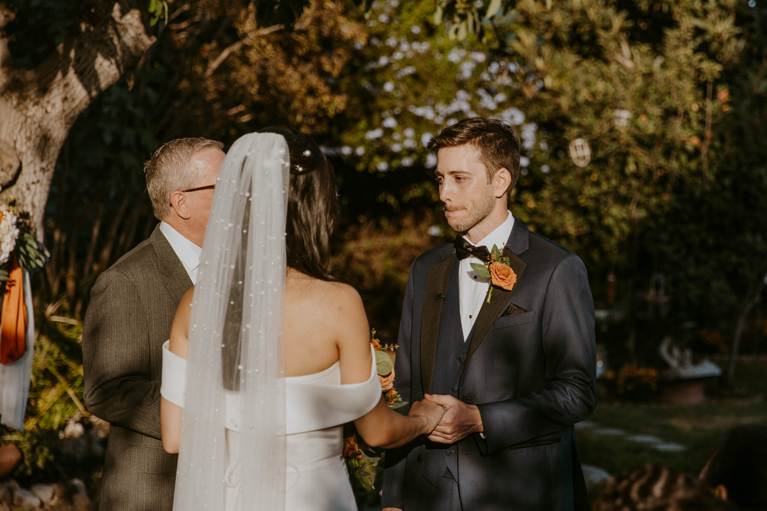 A bride and groom exchange vows at an outdoor wedding ceremony, with an officiant standing in the background at their intimate backyard wedding.