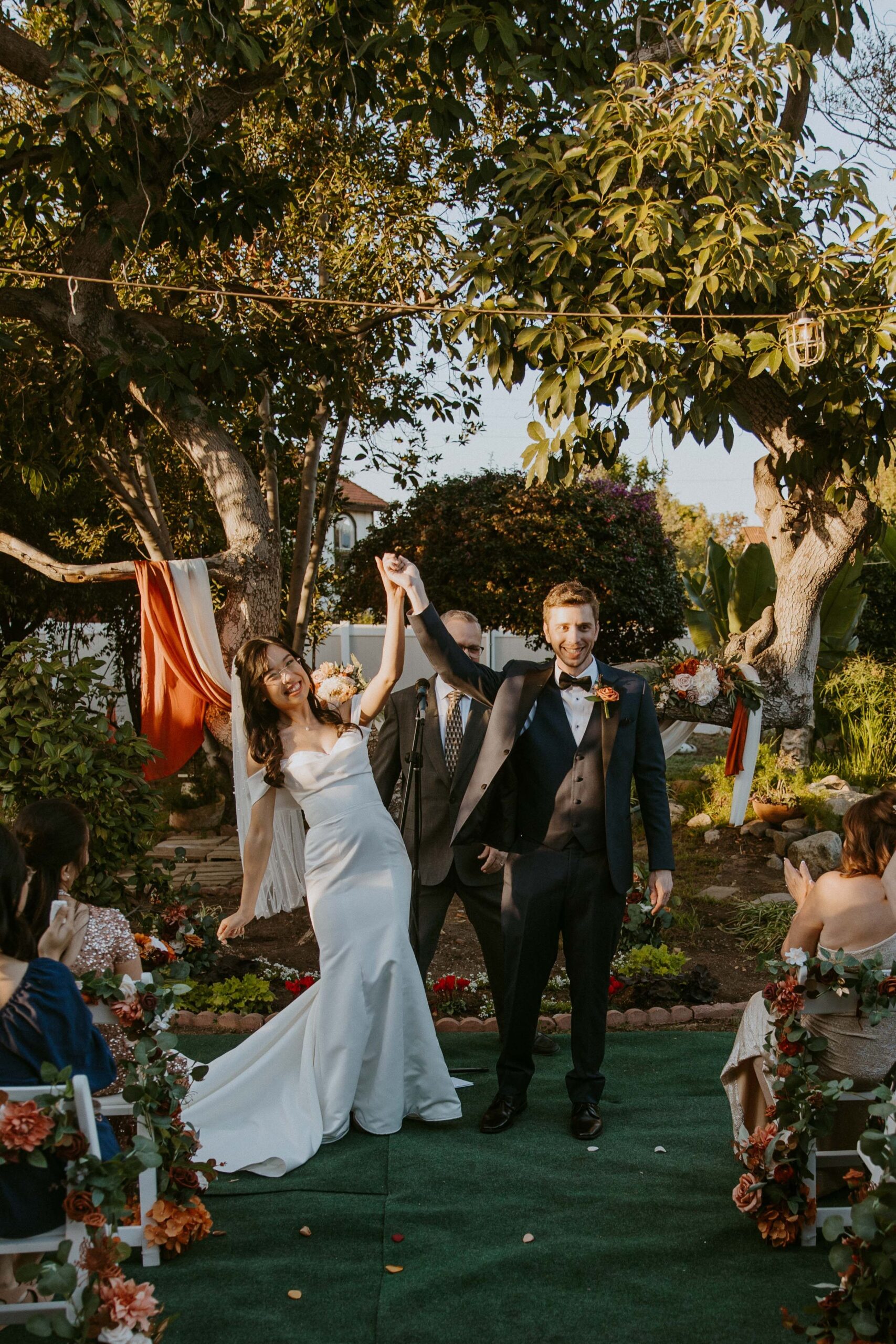 A newlywed couple walking down the aisle with their hands raised in celebration, surrounded by seated guests at their intimate backyard wedding. 