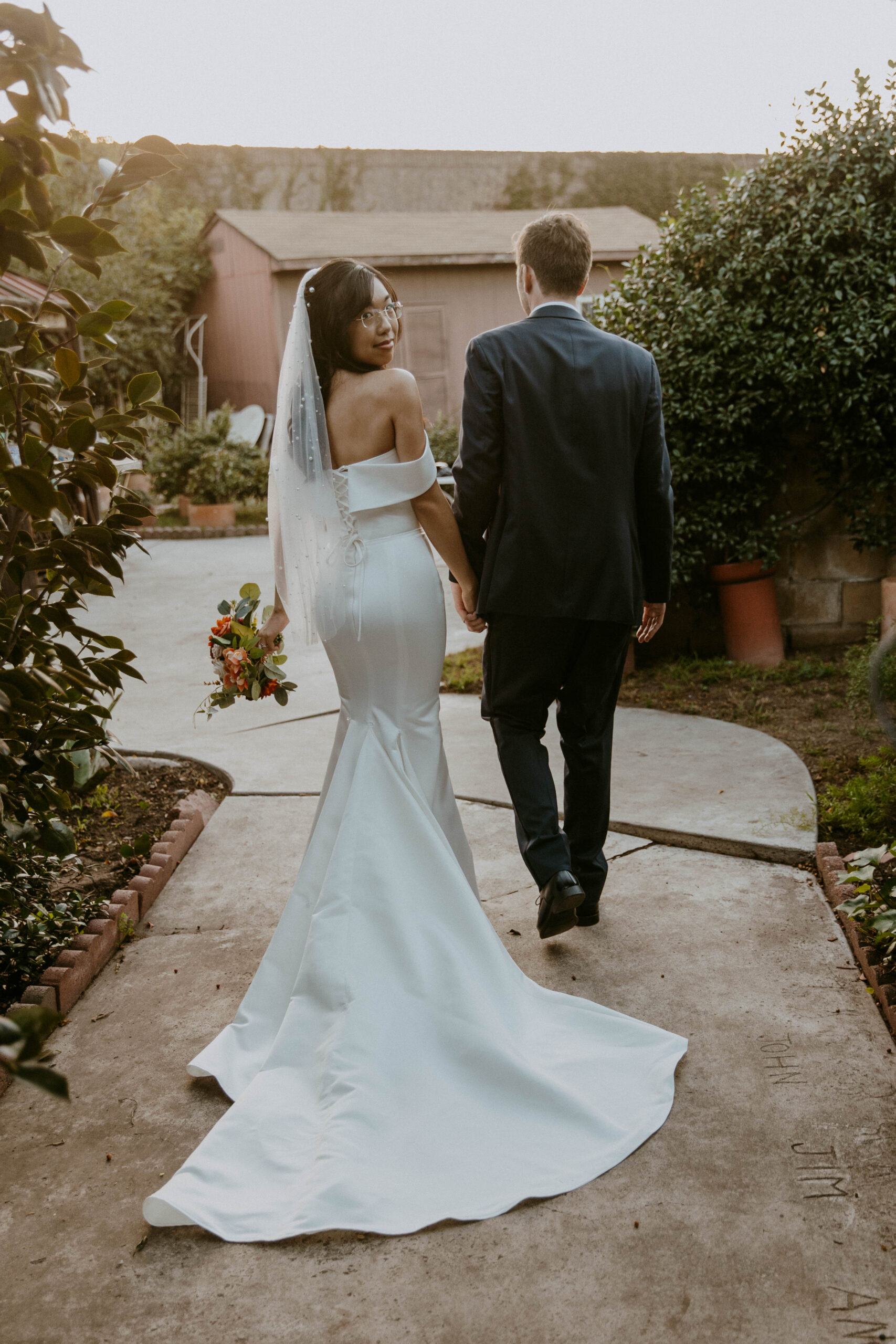 A bride in a white gown with a veil and a groom in a suit walking hand in hand away from the camera.