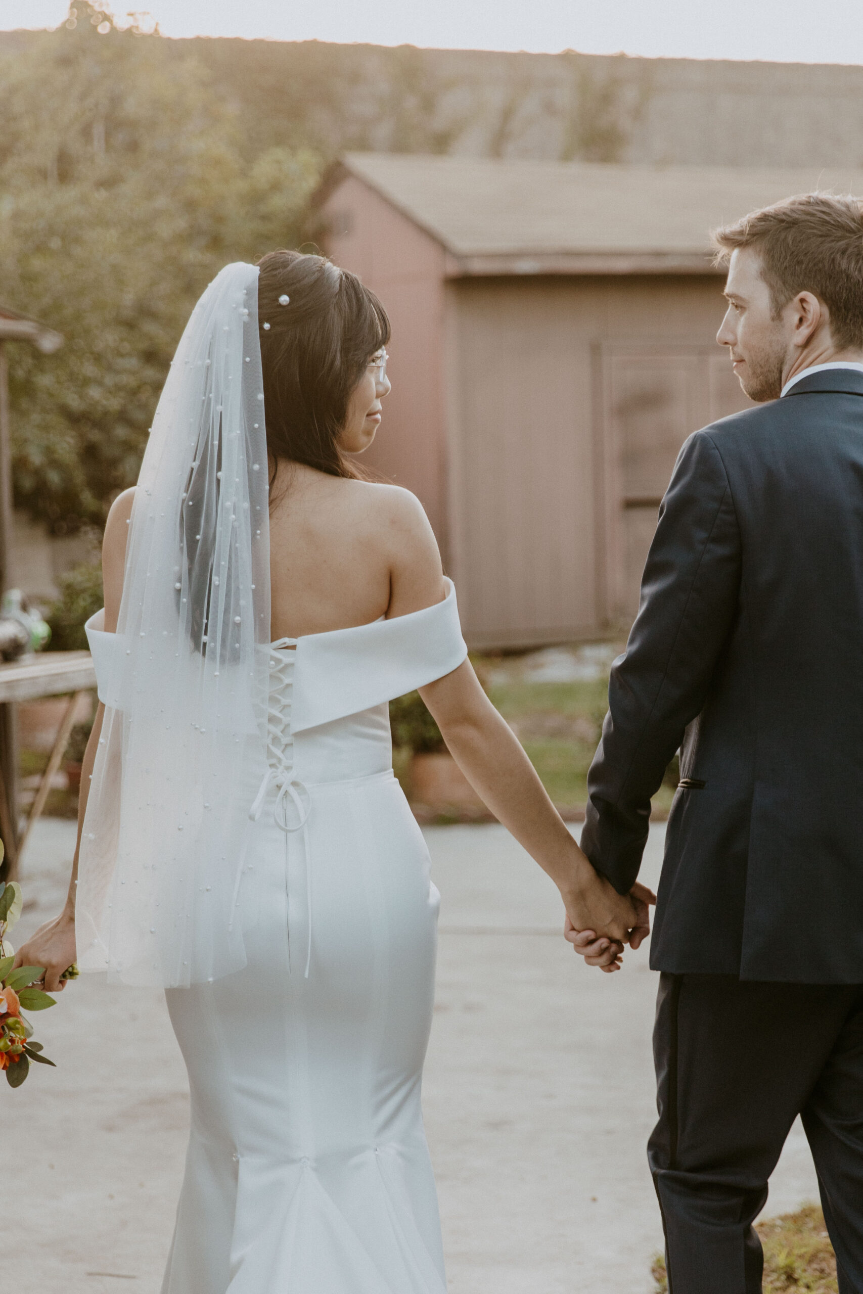 A bride in a white gown with a veil and a groom in a suit walking hand in hand away from the camera at their intimate backyard wedding