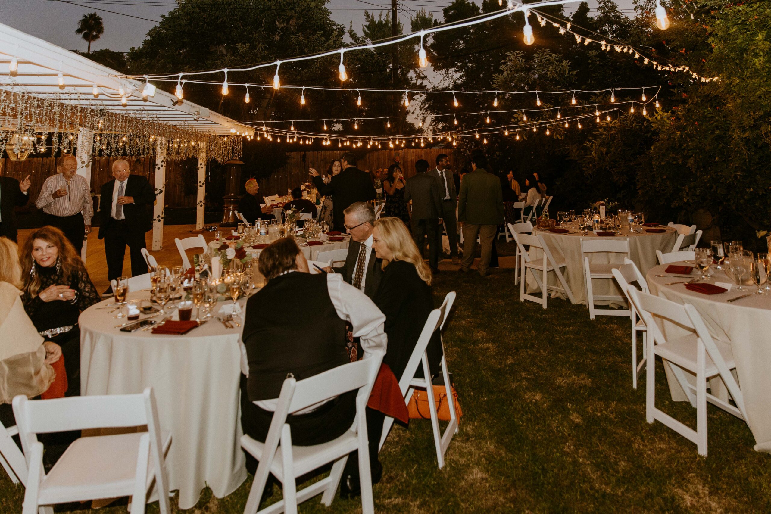 An evening outdoor intimate backyard wedding event with guests seated and mingling under string lights.