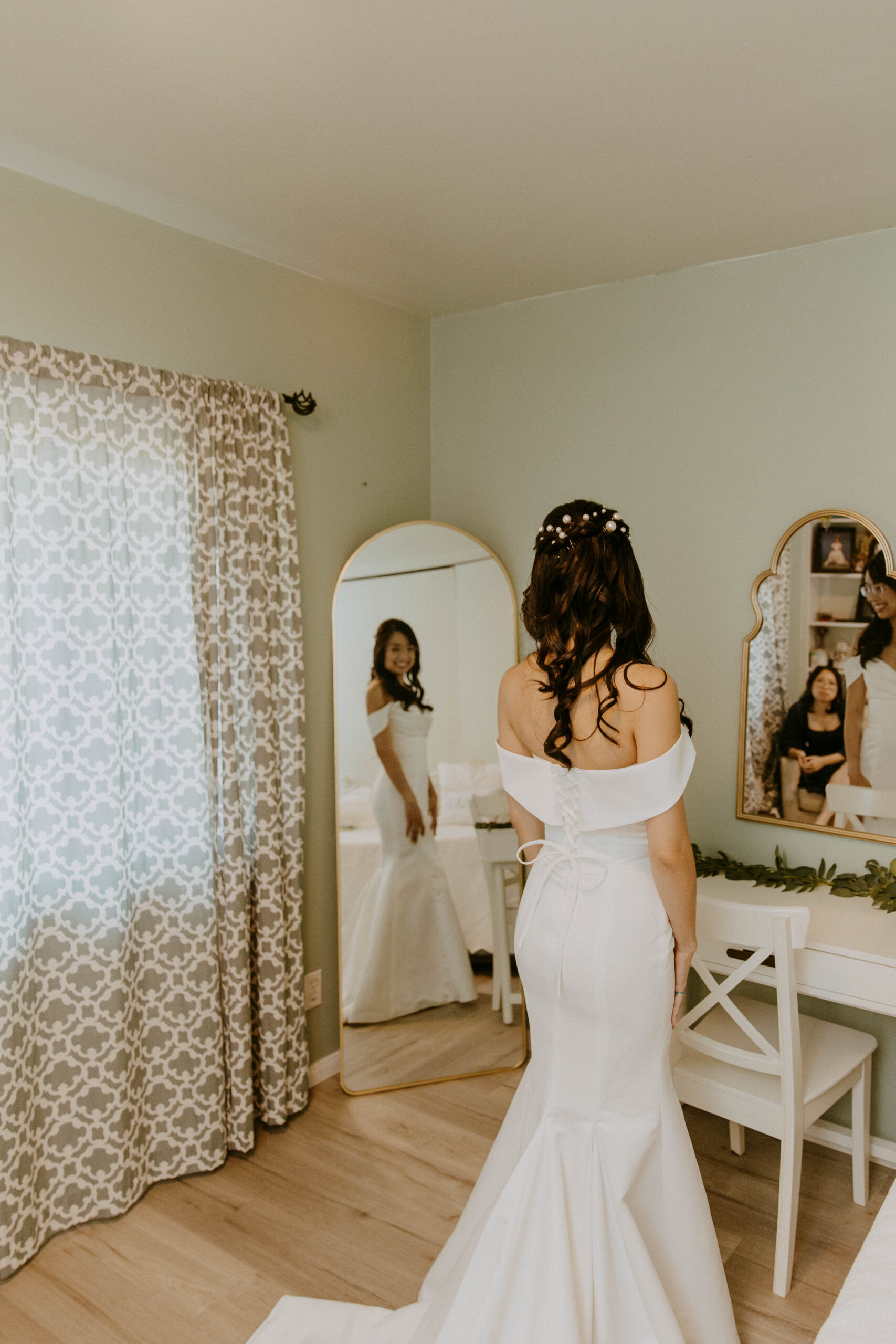 A woman in a white dress receives assistance with her wedding dress from another woman in a blue dress in front of a mirror.