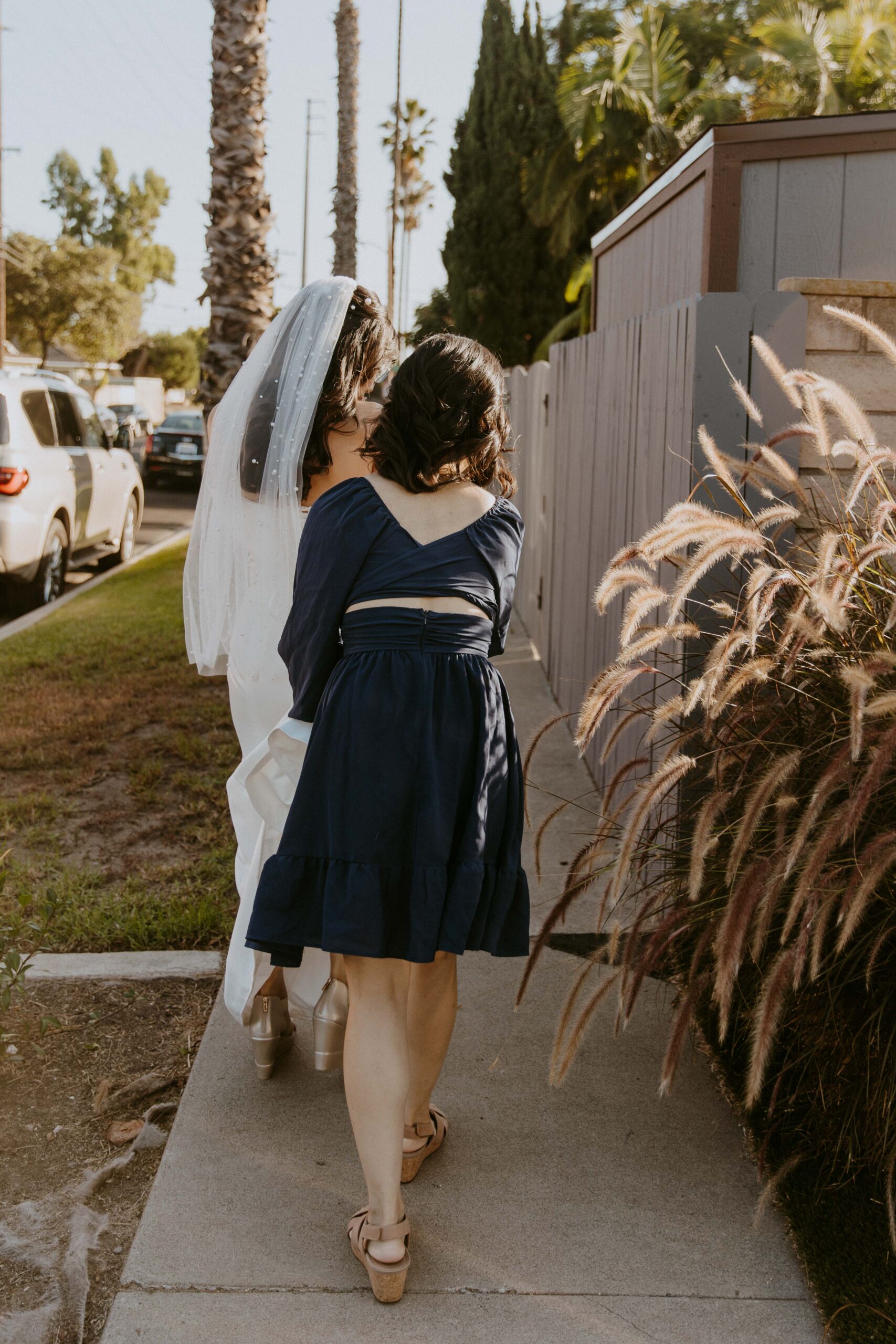 A bride and her attendant walking side by side on a sidewalk, with the bride carrying her veil and both women facing away from the camera.