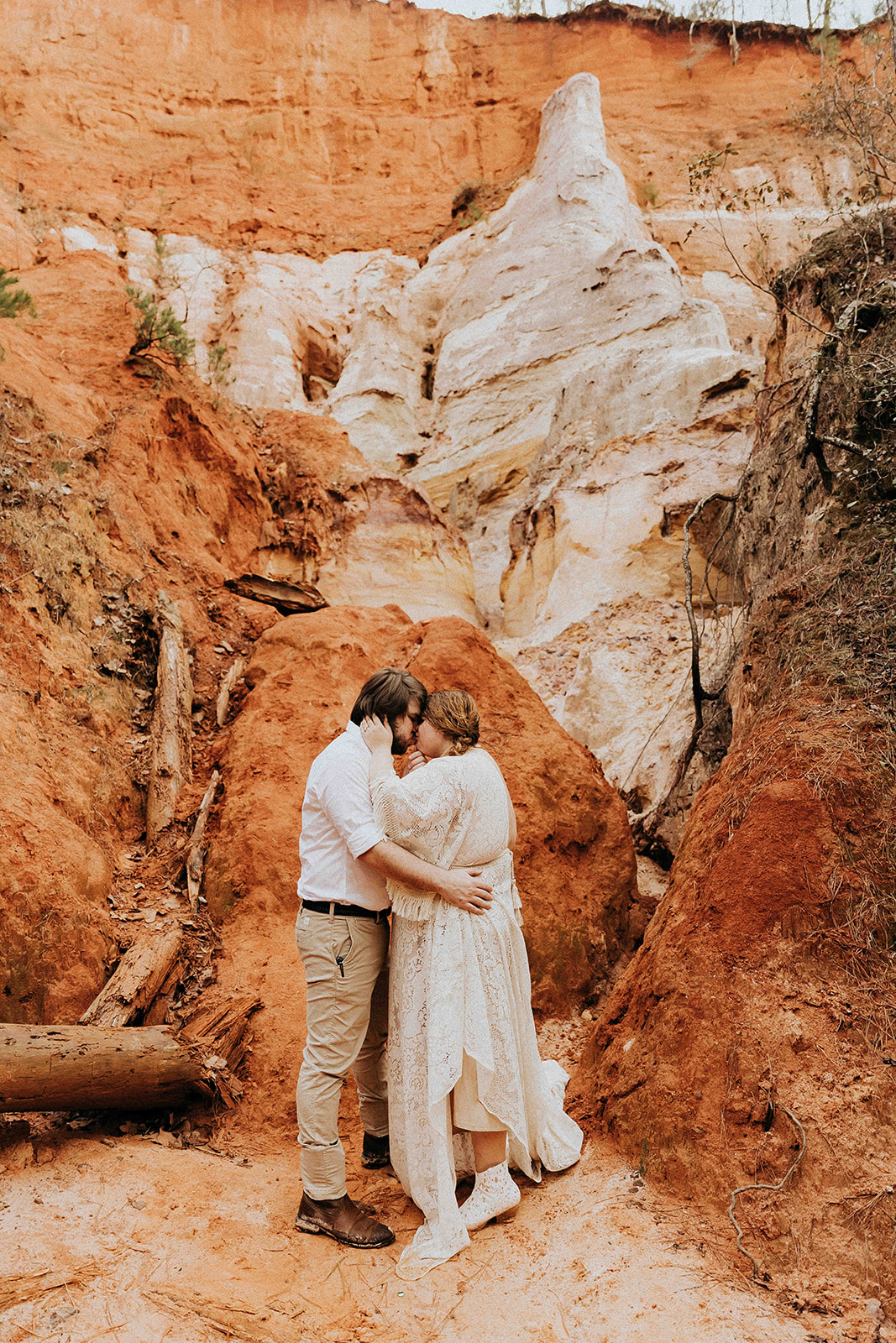 A couple stands back-to-back on a rocky landscape with reddish and white geological formations, surrounded by scattered fallen logs. The woman wears a long cream-colored dress and the man is in casual attire at a georgia elopement location