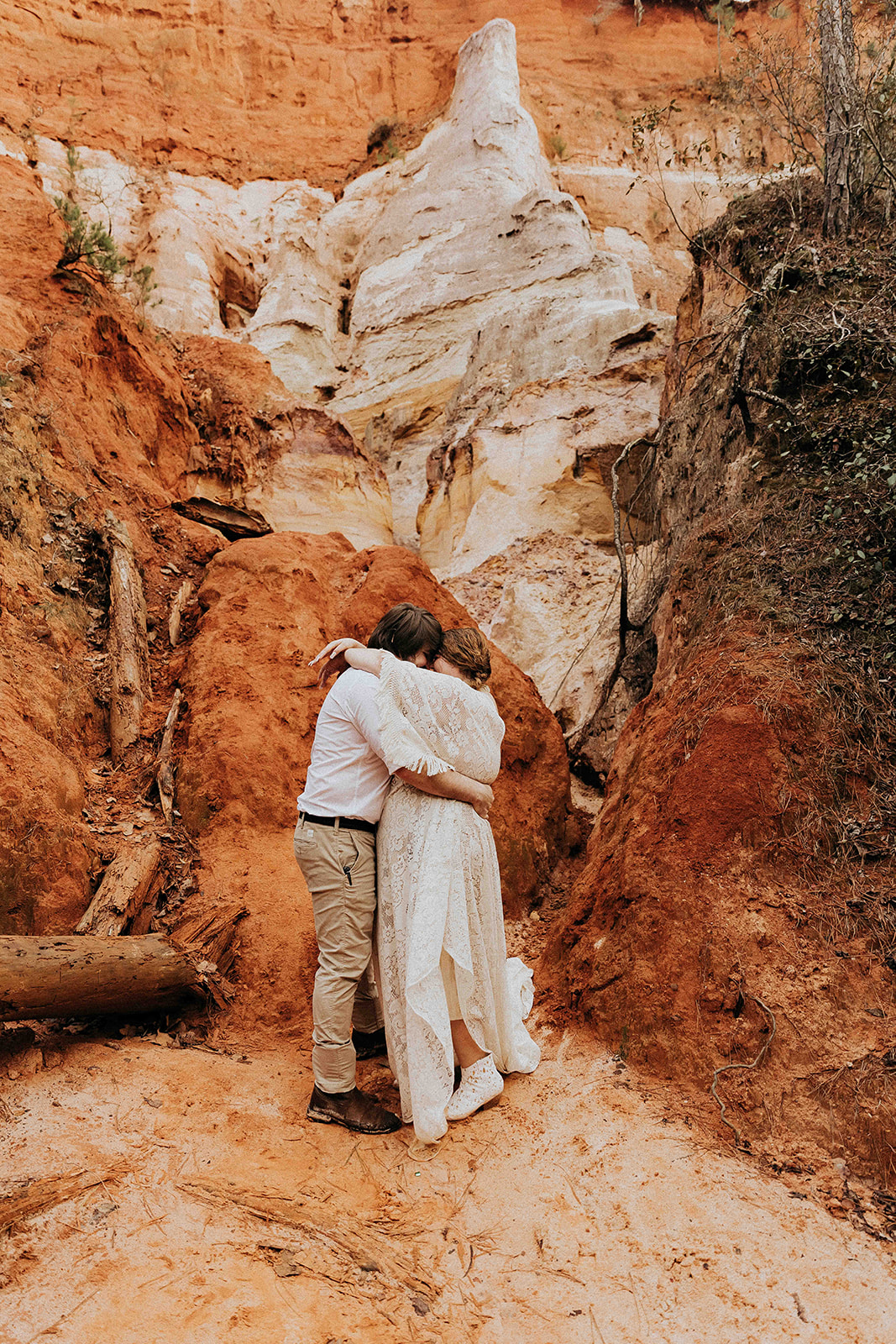 A couple stands back-to-back on a rocky landscape with reddish and white geological formations, surrounded by scattered fallen logs. The woman wears a long cream-colored dress and the man is in casual attire at a georgia elopement location