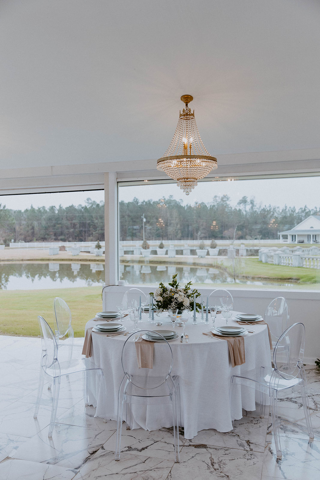 Elegant dining setup with a round table, ghost chairs, and a chandelier, overlooking a lake through large windows.