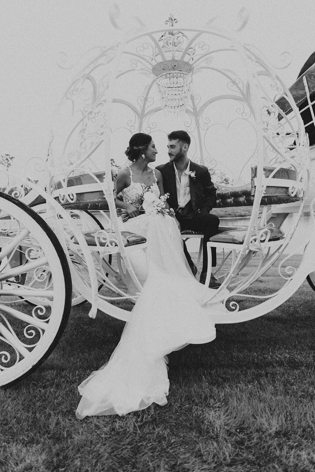 A bride and groom smiling and sitting inside an ornate white carriage with heart-shaped designs and a chandelier at Chateau 1800