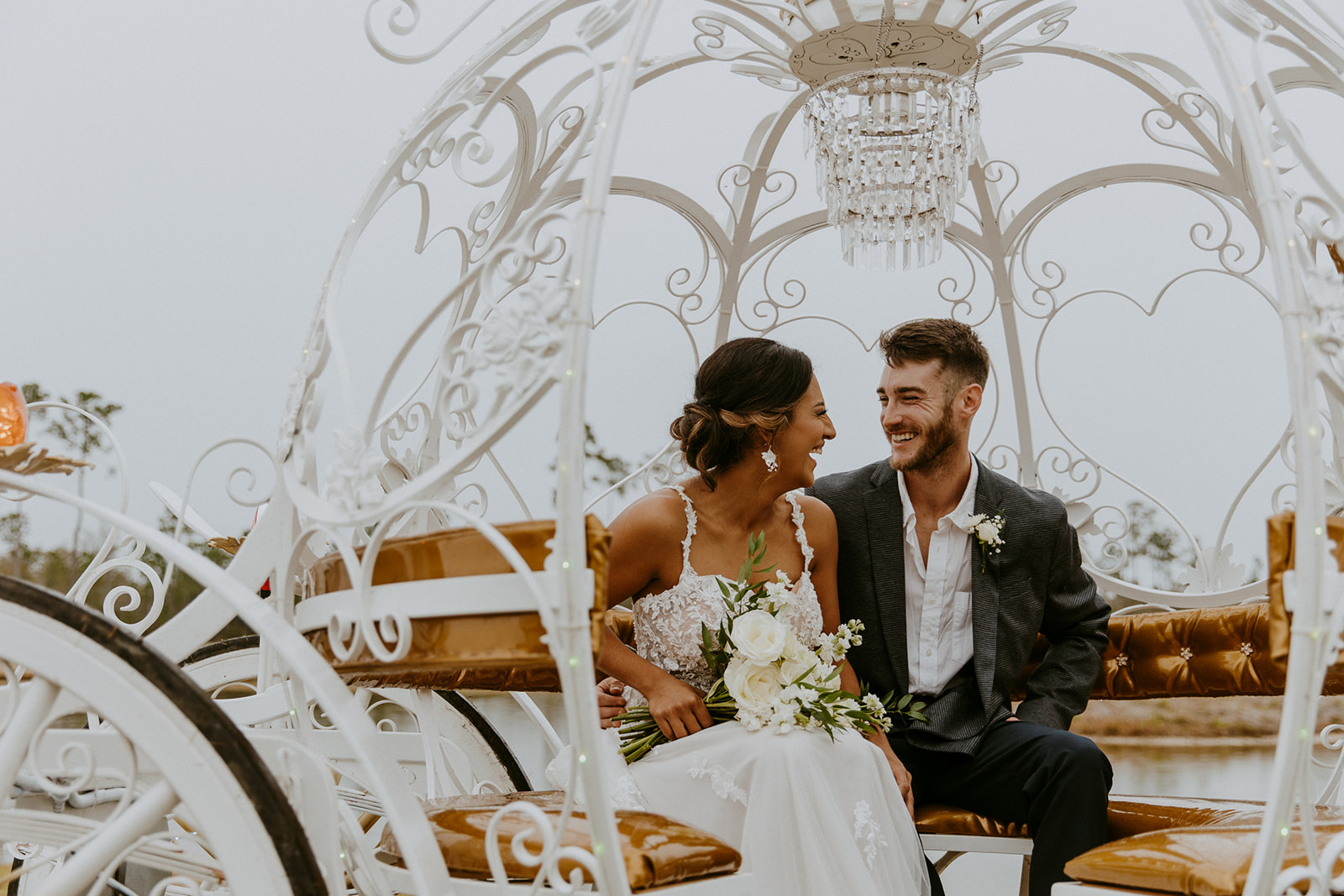 A bride and groom smiling and sitting inside an ornate white carriage with heart-shaped designs and a chandelier at Chateau 1800
