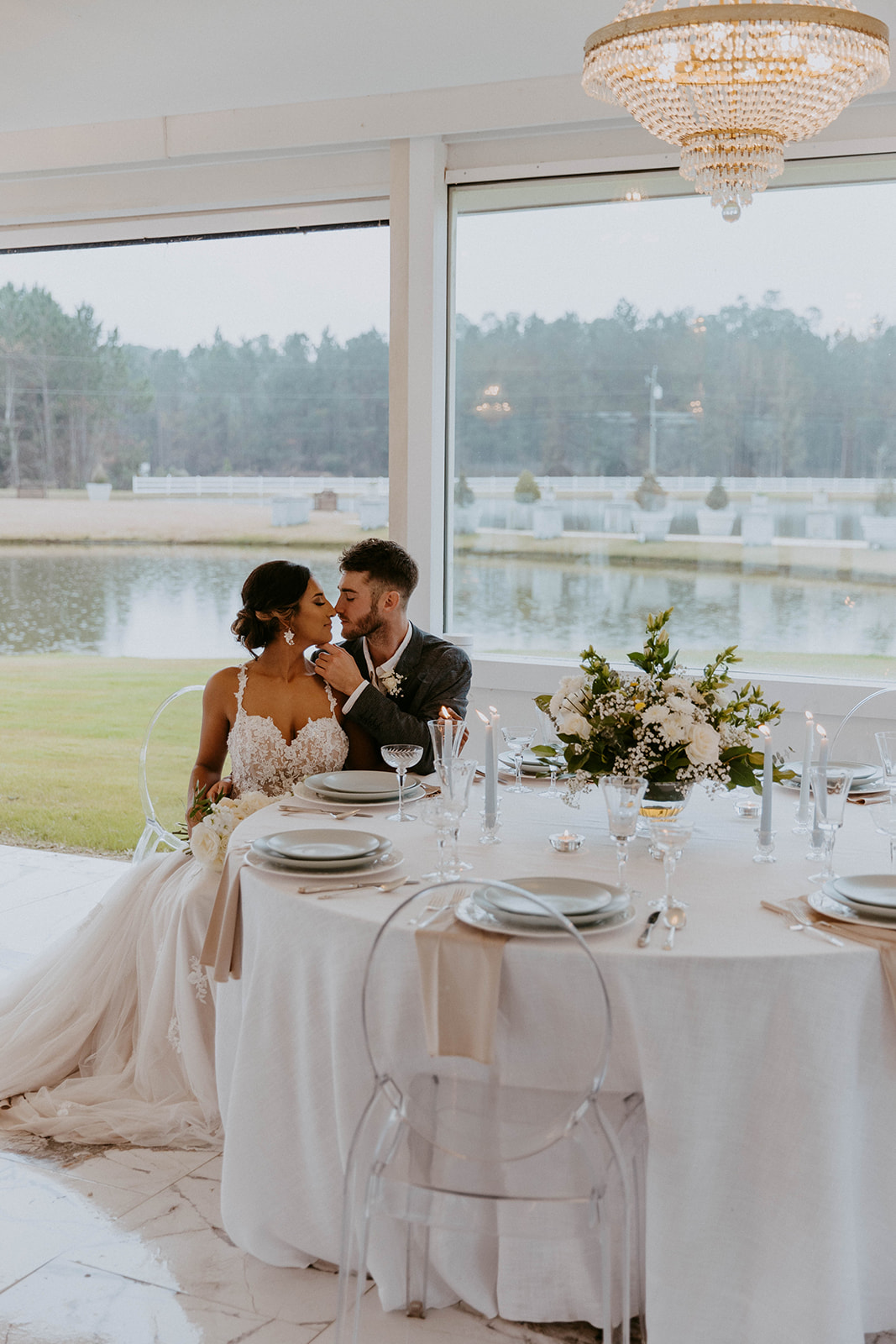 Bride and groom sitting at a dining table, sharing a kiss, with a scenic view of a lake through large windows in the background at Chateau 1800