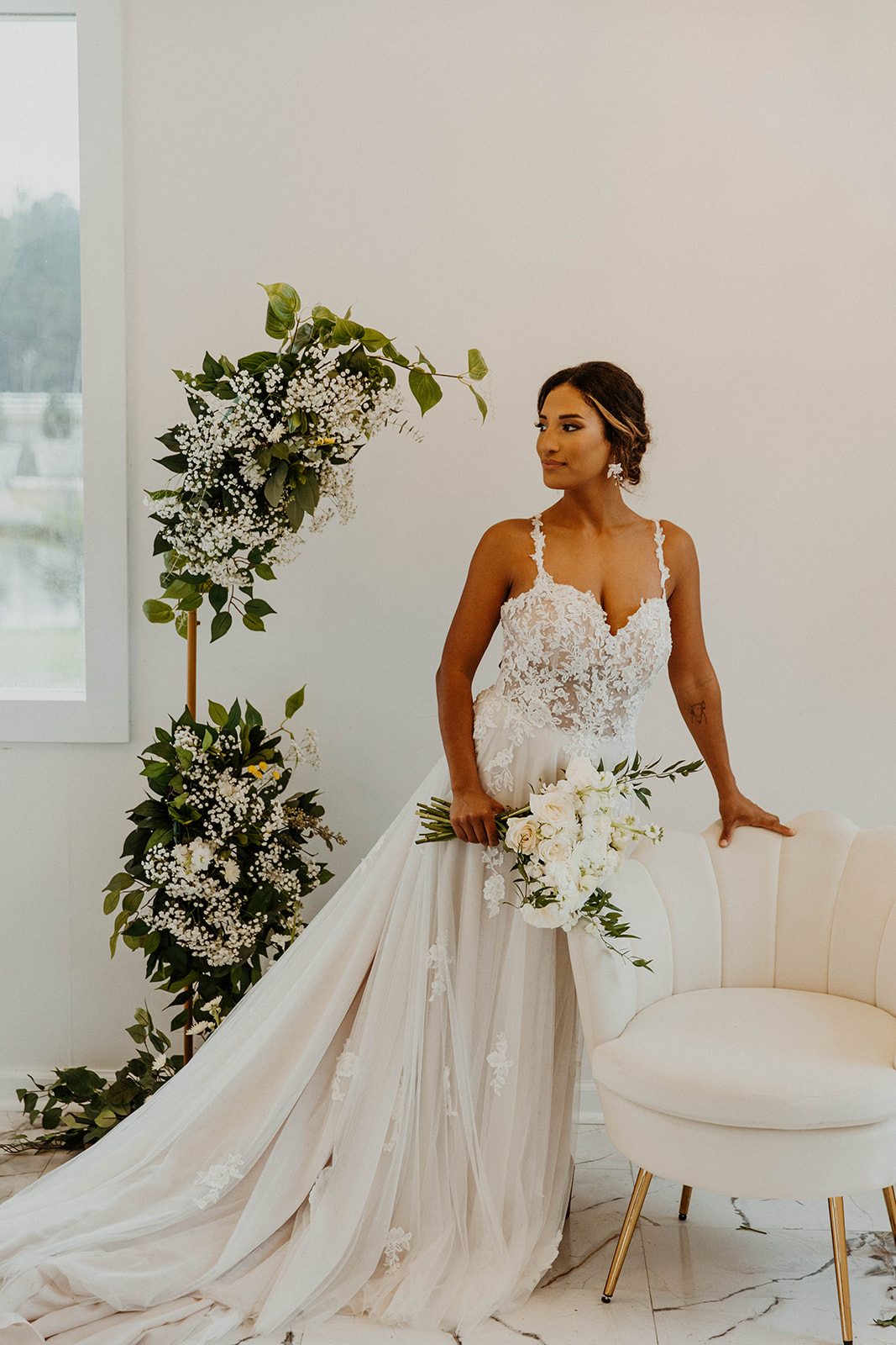A bride in a lace wedding dress poses with a bouquet beside a floral arrangement and a white sofa in a bright room atChateau 1800