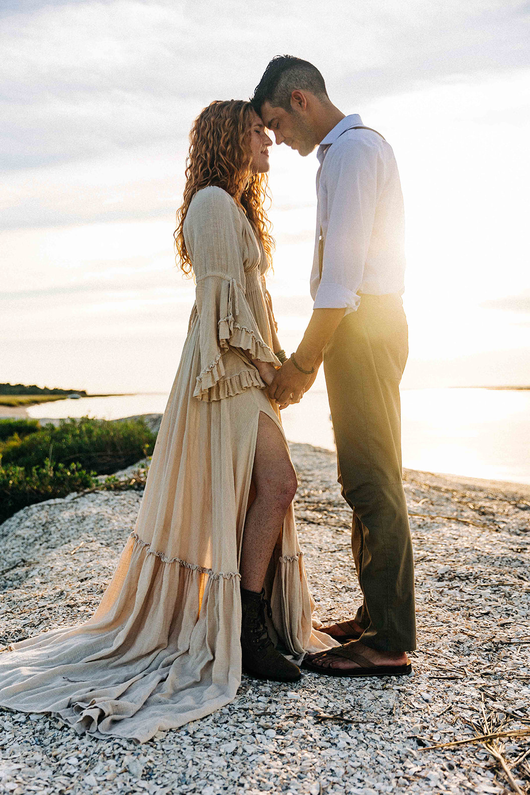 A couple in wedding attire walks on a beach at sunset, with the bride holding her gown and both smiling. Floral arrangements are scattered on the sand behind them at their georgia elopement location