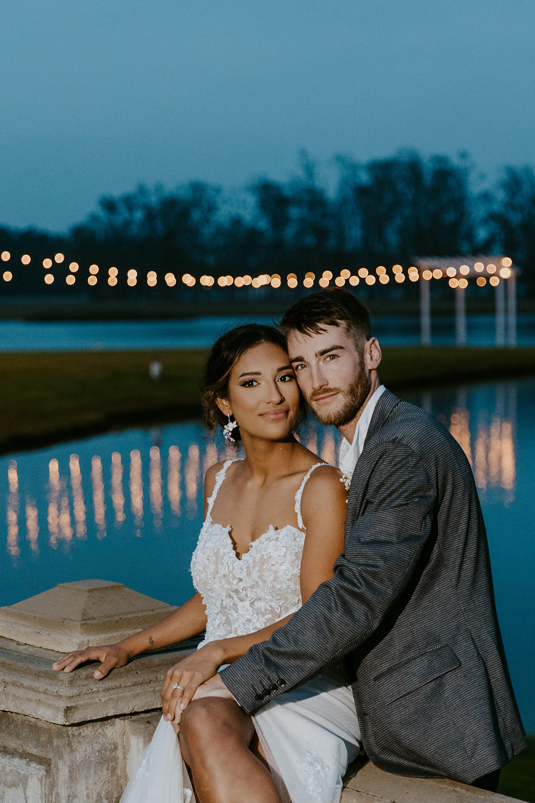 A newlywed couple poses at dusk, the woman in a white dress, the man in a gray suit, with a serene lake and string lights in the background.