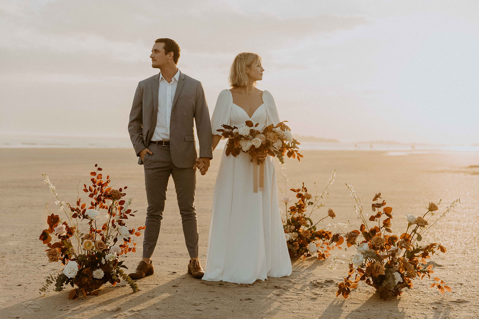A couple in wedding attire walks on a beach at sunset, with the bride holding her gown and both smiling. Floral arrangements are scattered on the sand behind them at their georgia elopement location