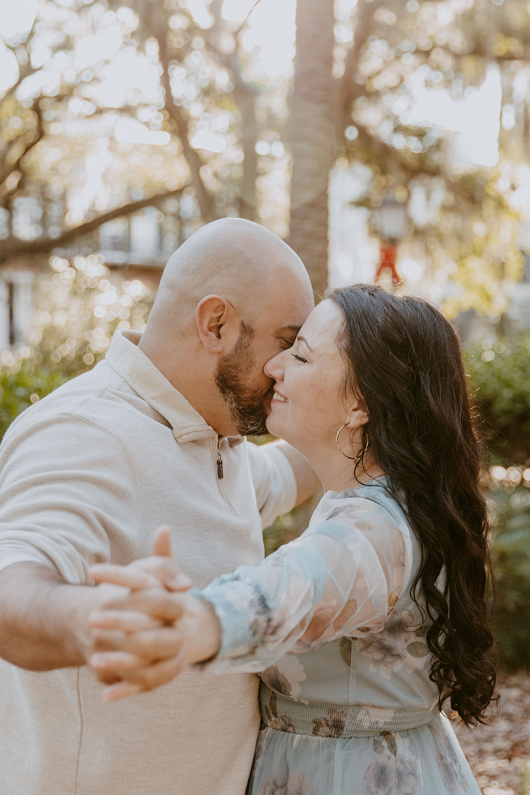 A man and a woman with long dark hair outdoors in a garden-like setting. Both have their eyes closed, showing a moment of affection in Downtown Historic district savannah ga