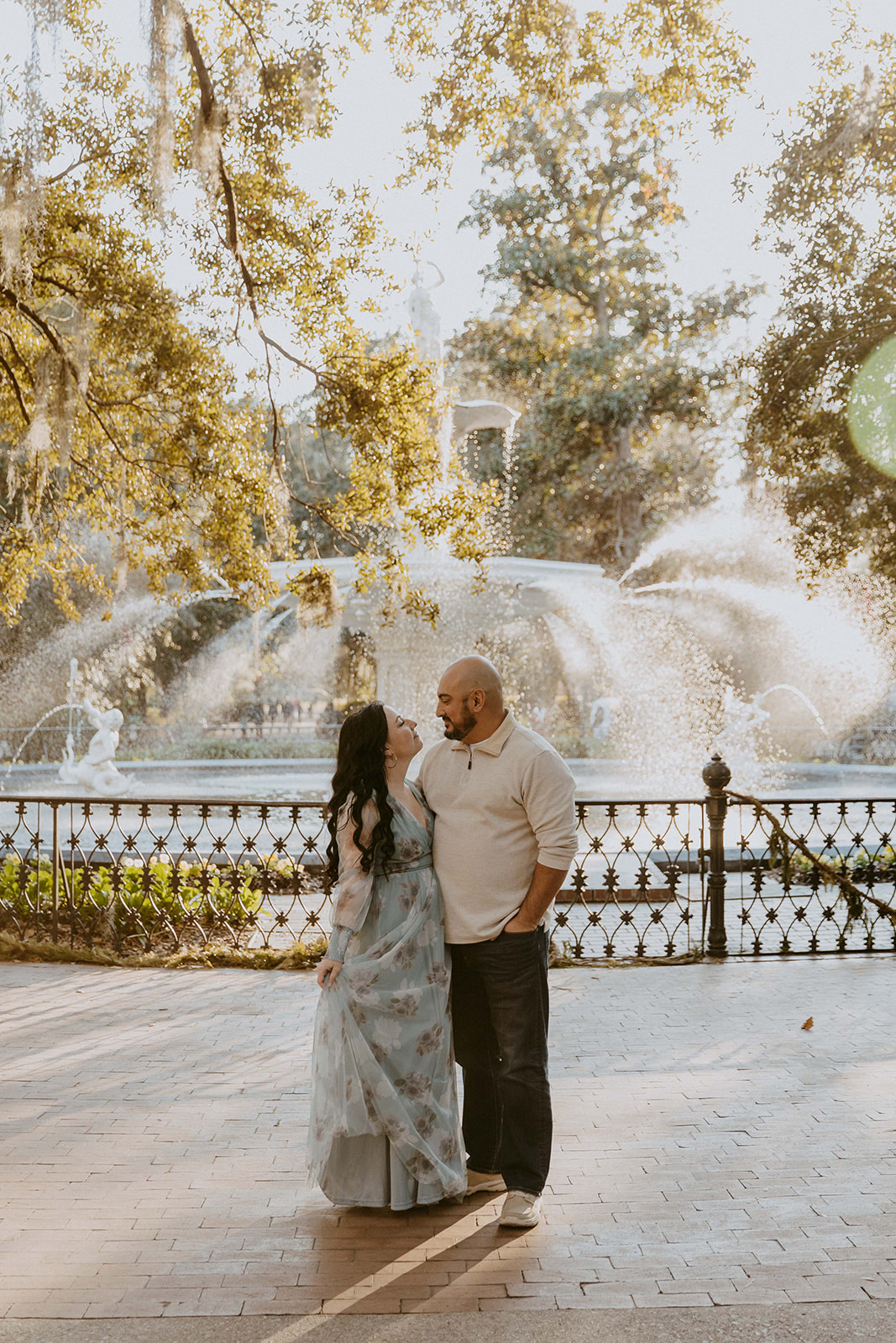 A man and a woman with long dark hair outdoors in a garden-like setting during their engagement session