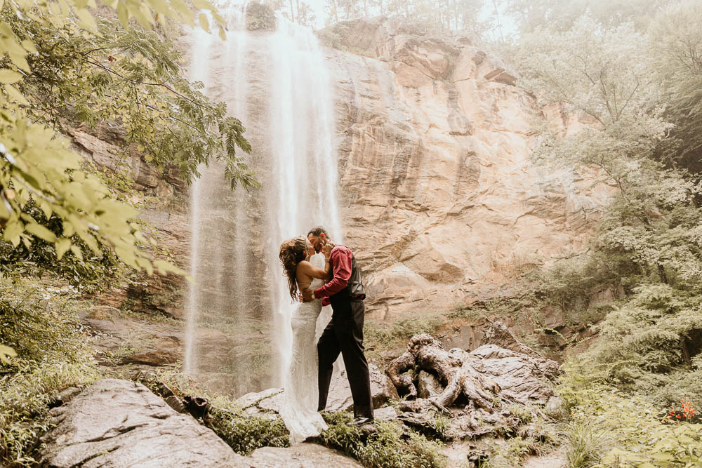 A couple embraces in front of a waterfall. The man is wearing a red shirt with a black vest, and the woman is in a white dress during their georgia elopement location taccoa falls