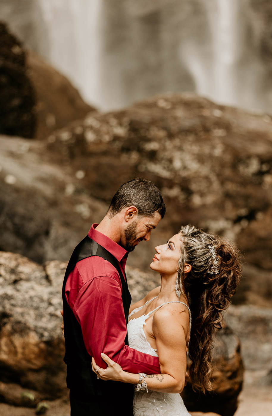 A couple embraces in front of a waterfall. The man is wearing a red shirt with a black vest, and the woman is in a white dress during their georgia elopement location taccoa falls