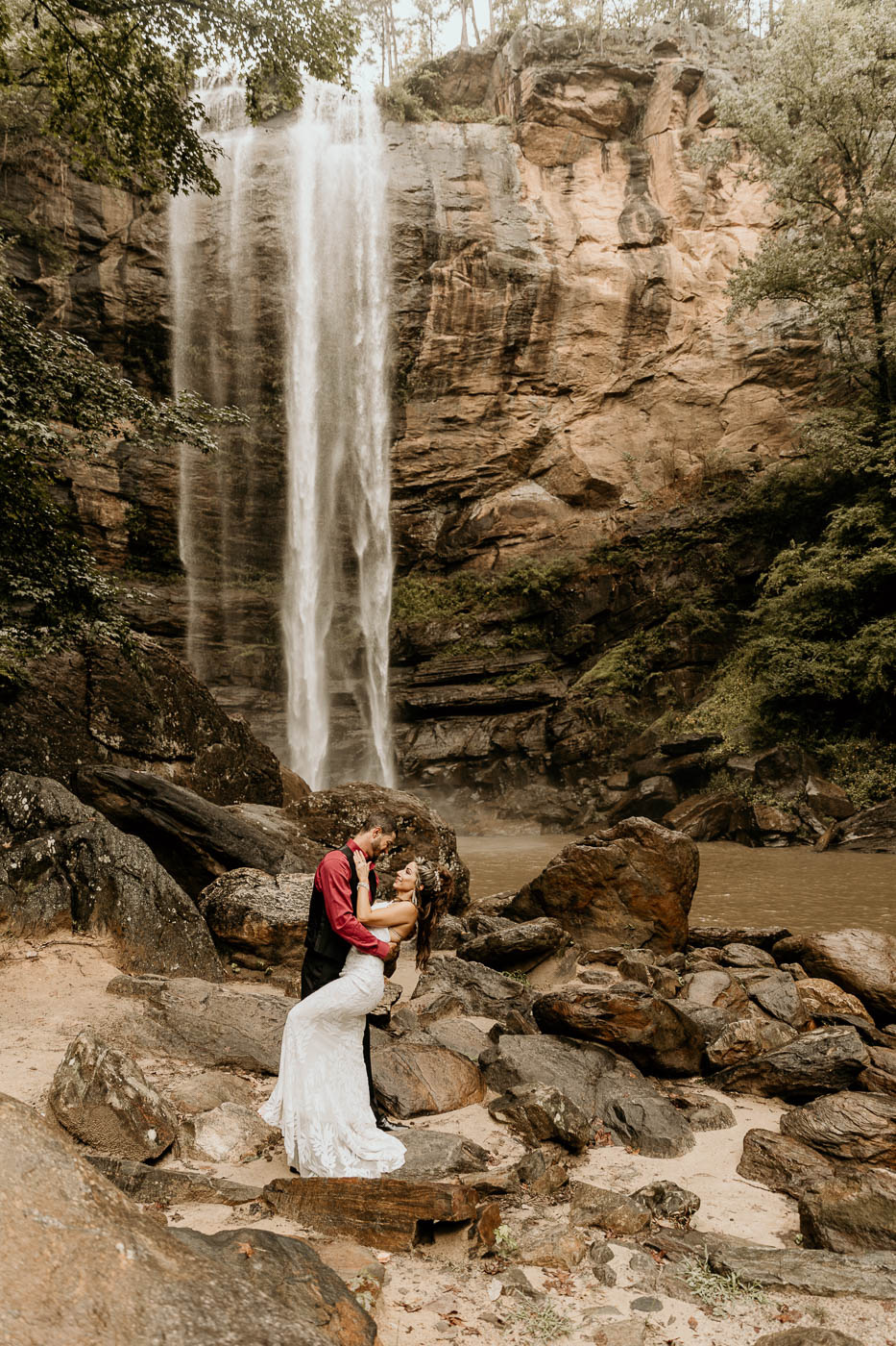 A couple embraces in front of a waterfall. The man is wearing a red shirt with a black vest, and the woman is in a white dress at a georgia elopement location taccoa falls