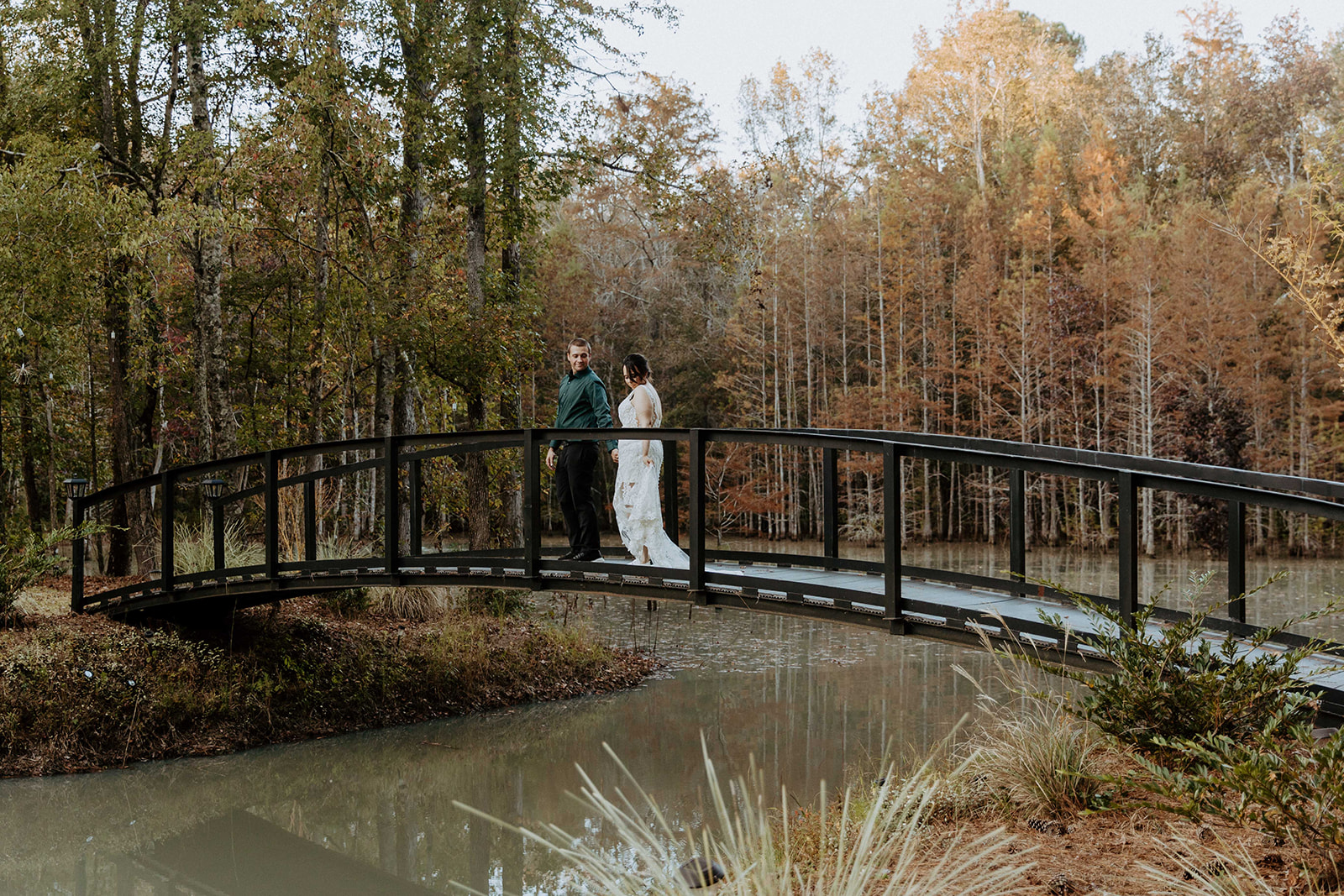 A bride in a lace dress and a groom in a black suit stand close together on a leaf-covered dock by a lake with trees in the background. The bride holds a bouquet of flowers.