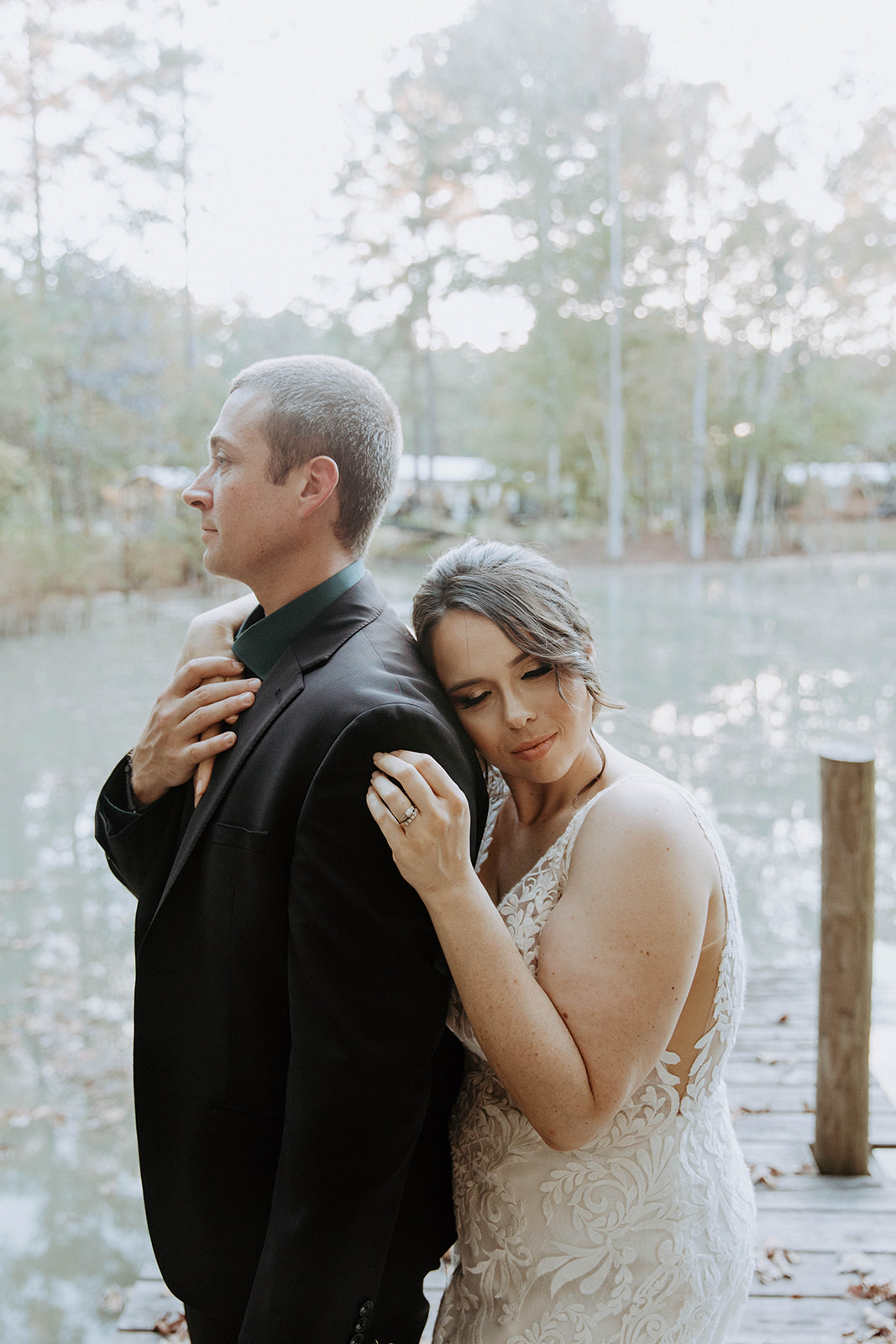 A bride in a lace dress and a groom in a black suit stand close together on a leaf-covered dock by a lake with trees in the background. The bride holds a bouquet of flowers.