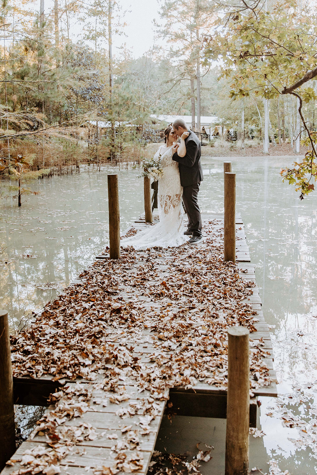 A newlywed couple stands outdoors, the groom in a black suit holding the bride in a white dress who is holding a bouquet. The setting is grassy with trees in the background at their fall elopement