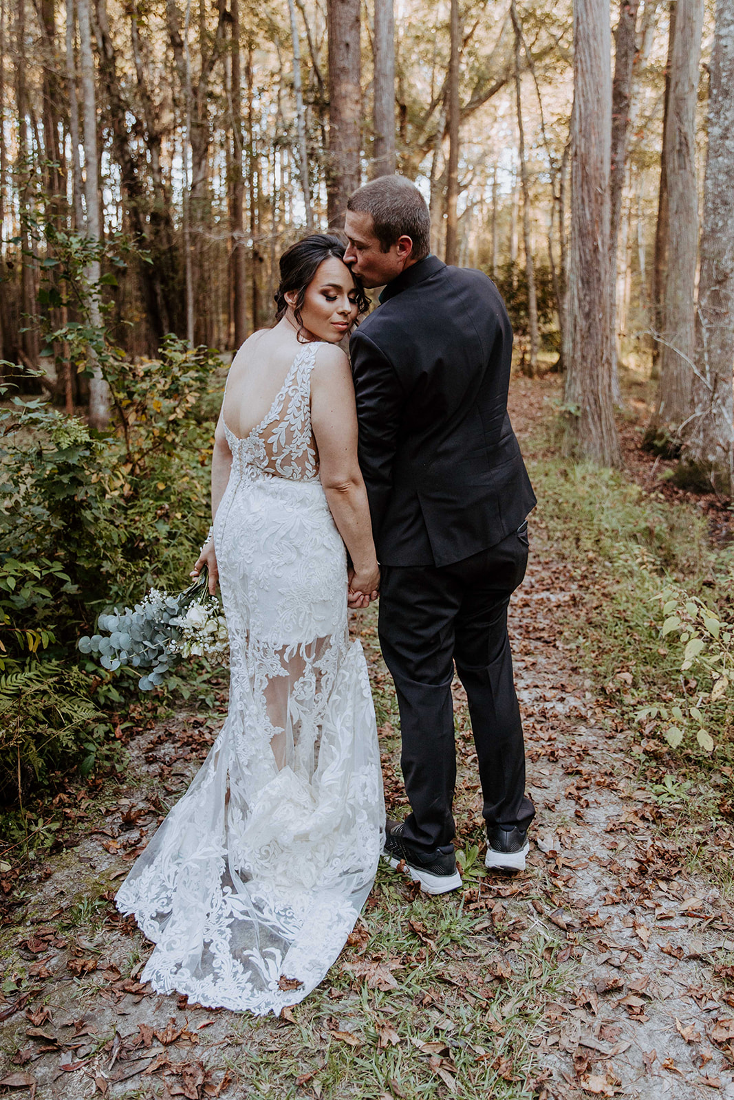 A newlywed couple stands outdoors, the groom in a black suit holding the bride in a white dress who is holding a bouquet. The setting is grassy with trees in the background at their fall elopement