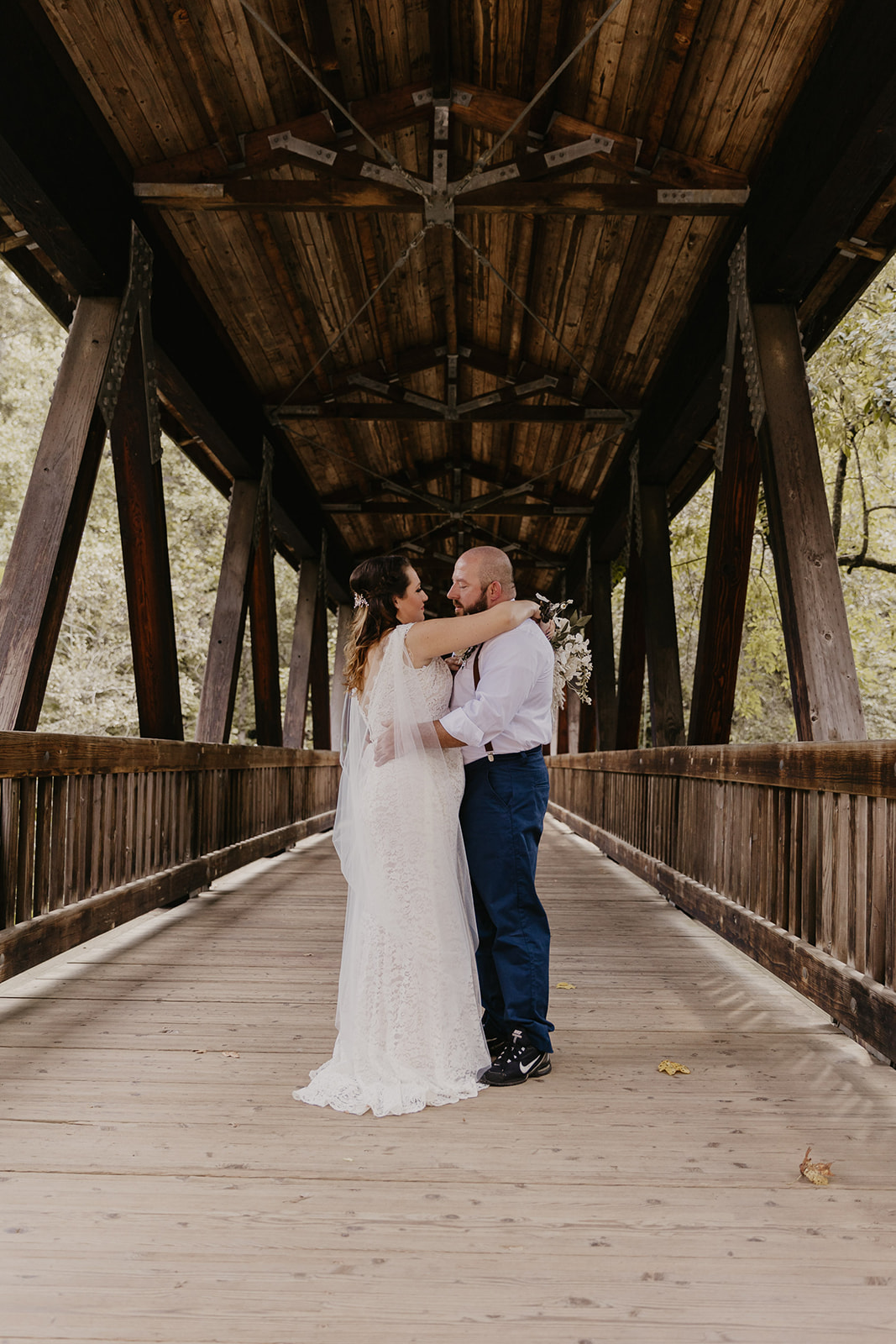 A couple, dressed in wedding attire, embraces on a wooden covered bridge with a backdrop of trees.