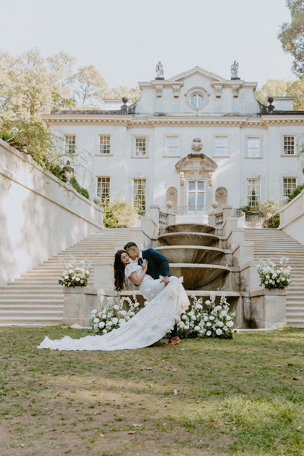 A couple in formal attire walks up the steps of a large, elegant building with a well-manicured garden and fountains 