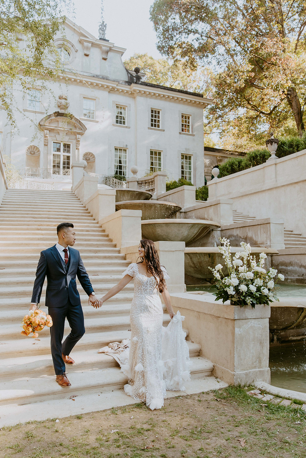 A couple in formal attire walks up the steps of a large, elegant building with a well-manicured garden and fountains 