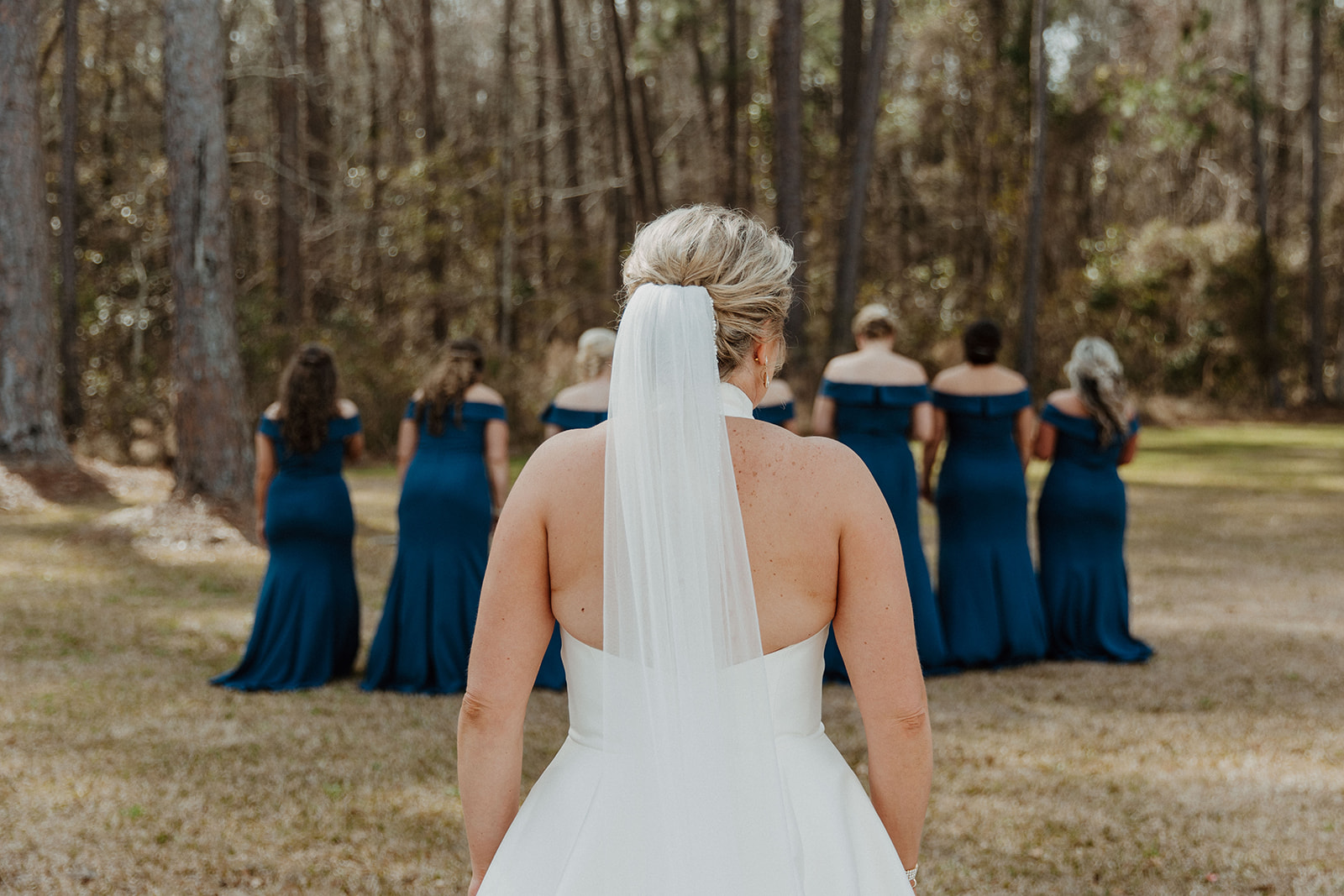 A bride in a white dress with a veil stands facing a group of bridesmaids in dark blue dresses, who are turned away, in a wooded outdoor setting during a first look with her bridesmaid at The Venues at Ogeechee Tech
