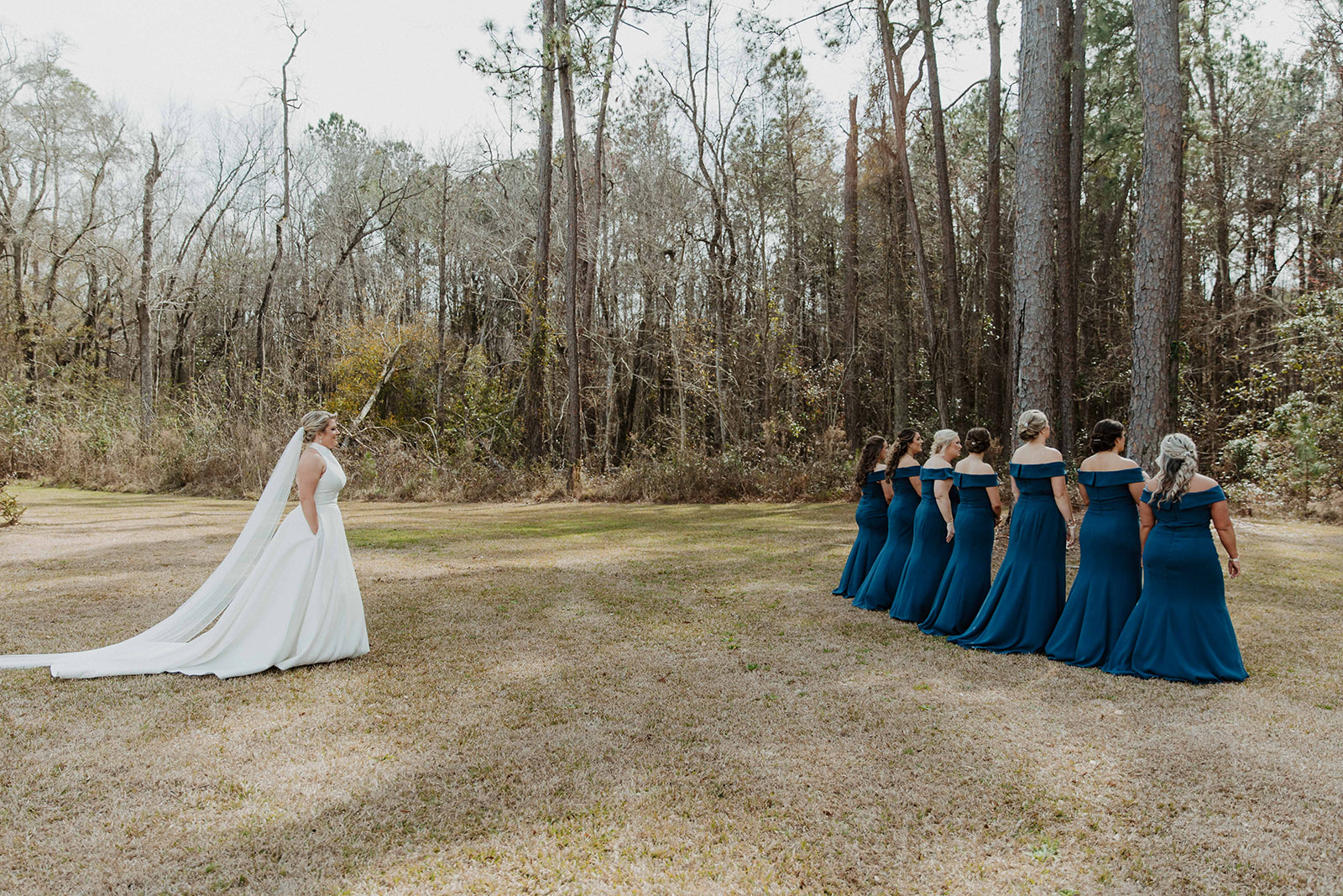 A bride in a white dress with a veil stands facing a group of bridesmaids in dark blue dresses, who are turned away, in a wooded outdoor setting during a first look with her bridesmaid at The Venues at Ogeechee Tech