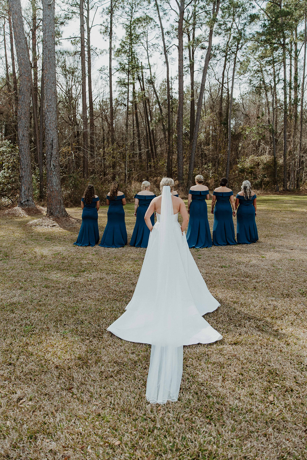 A bride in a white dress with a veil stands facing a group of bridesmaids in dark blue dresses, who are turned away, in a wooded outdoor setting during a first look with her bridesmaid at The Venues at Ogeechee Tech