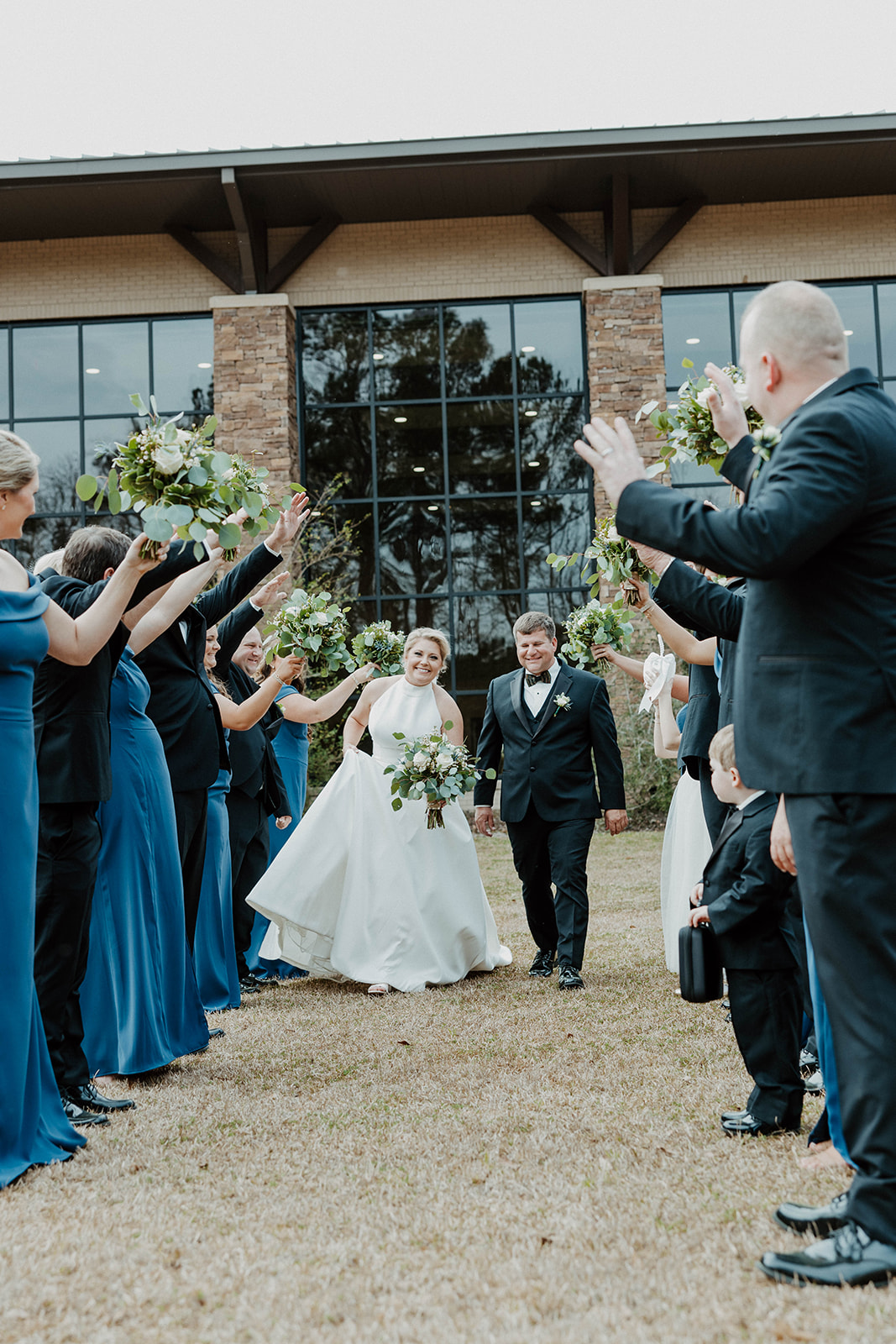 A newlywed couple walks under a tunnel of raised hands holding bouquets, with bridesmaids in blue dresses and groomsmen in dark suits standing on either side.