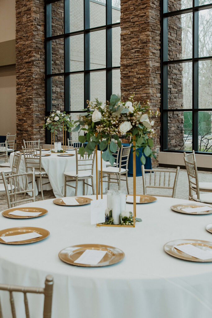 A round table set for an event with white tablecloth, gold-rimmed plates, and a tall floral centerpiece. The backdrop includes large windows and stone columns.