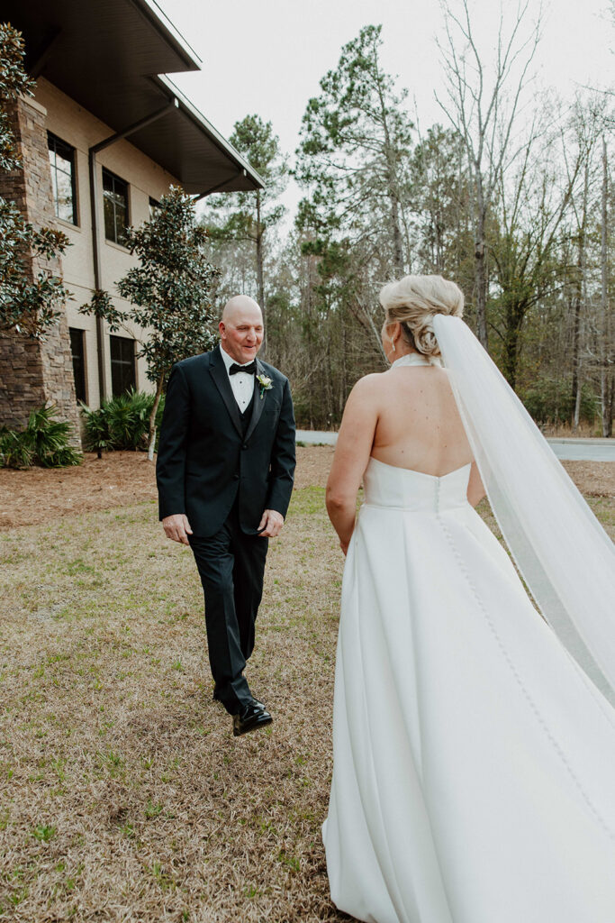 A bride in a white gown with a long train walks towards a man in a suit outside a large building with stone accents and tall windows during a first look with dad at The Venues at Ogeechee Tech