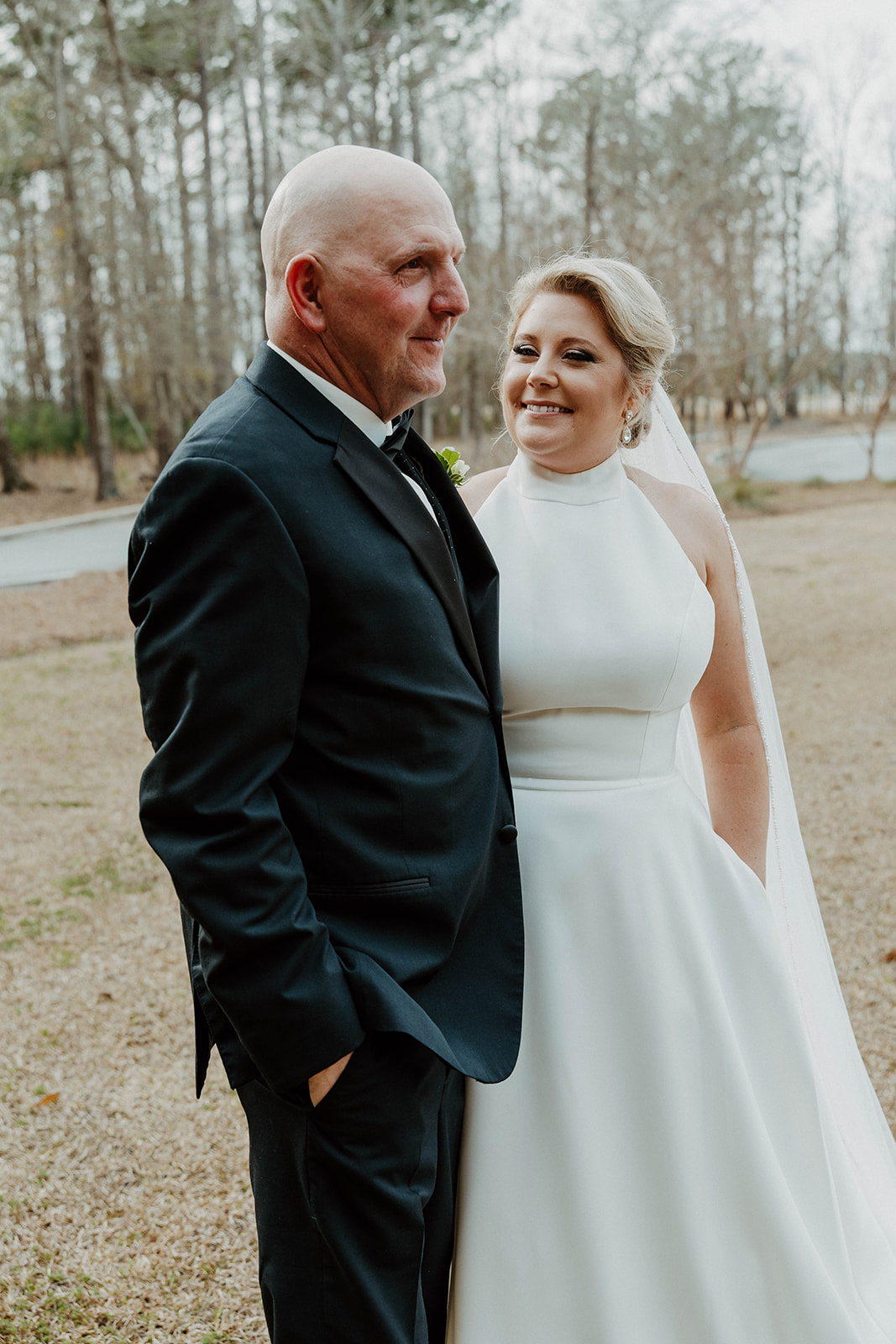 A bride in a white gown with a long train walks towards a man in a suit outside a large building with stone accents and tall windows during a first look with dad at The Venues at Ogeechee Tech