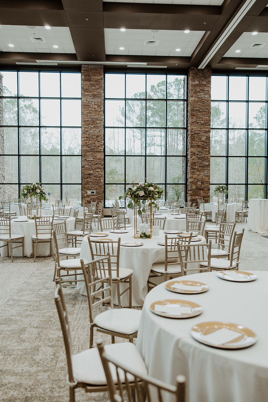 A round table set for an event with white tablecloth, gold-rimmed plates, and a tall floral centerpiece. The backdrop includes large windows and stone columns.