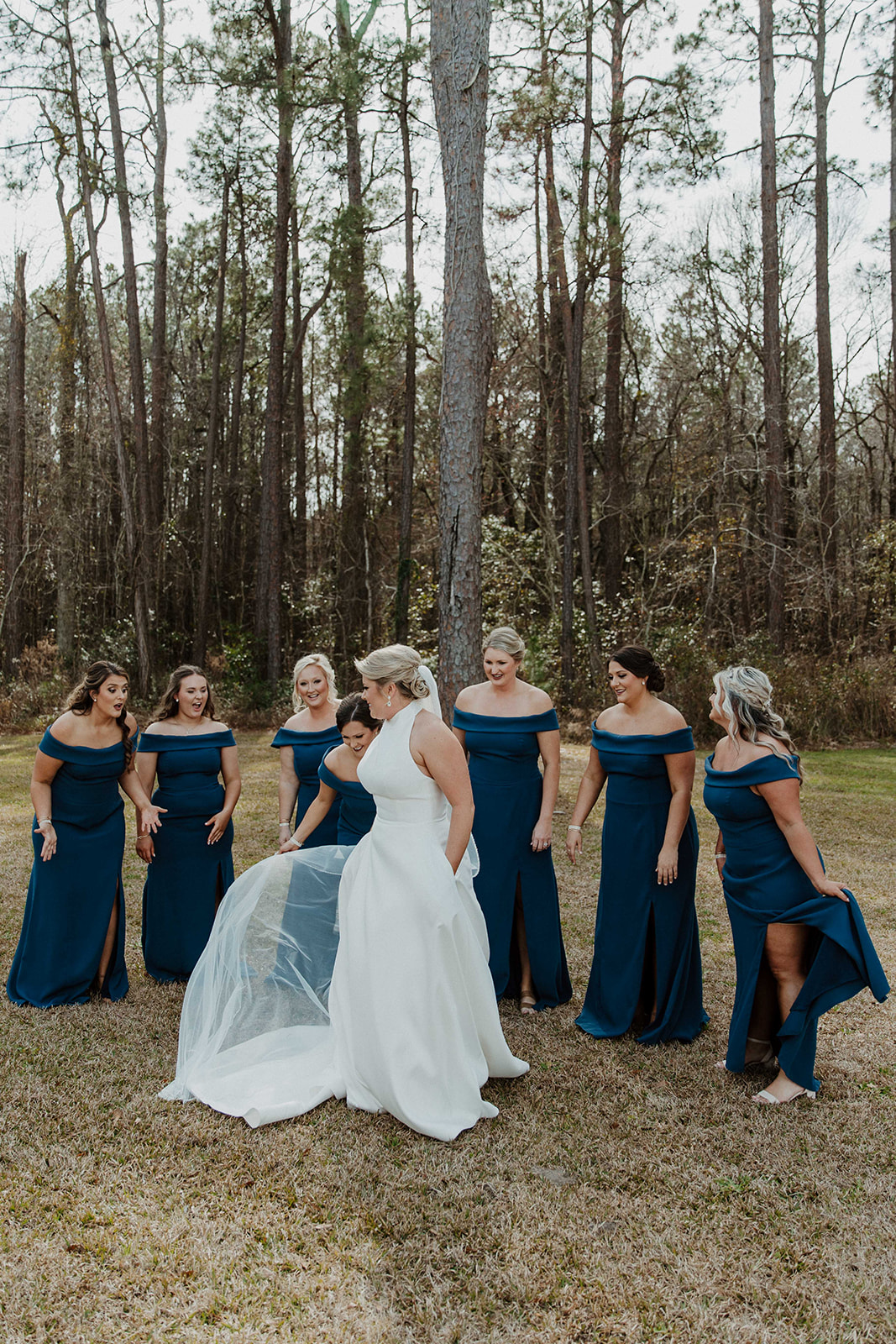 A bride in a white dress with a veil stands facing a group of bridesmaids in dark blue dresses, who are turned away, in a wooded outdoor setting during a first look with her bridesmaid at The Venues at Ogeechee Tech