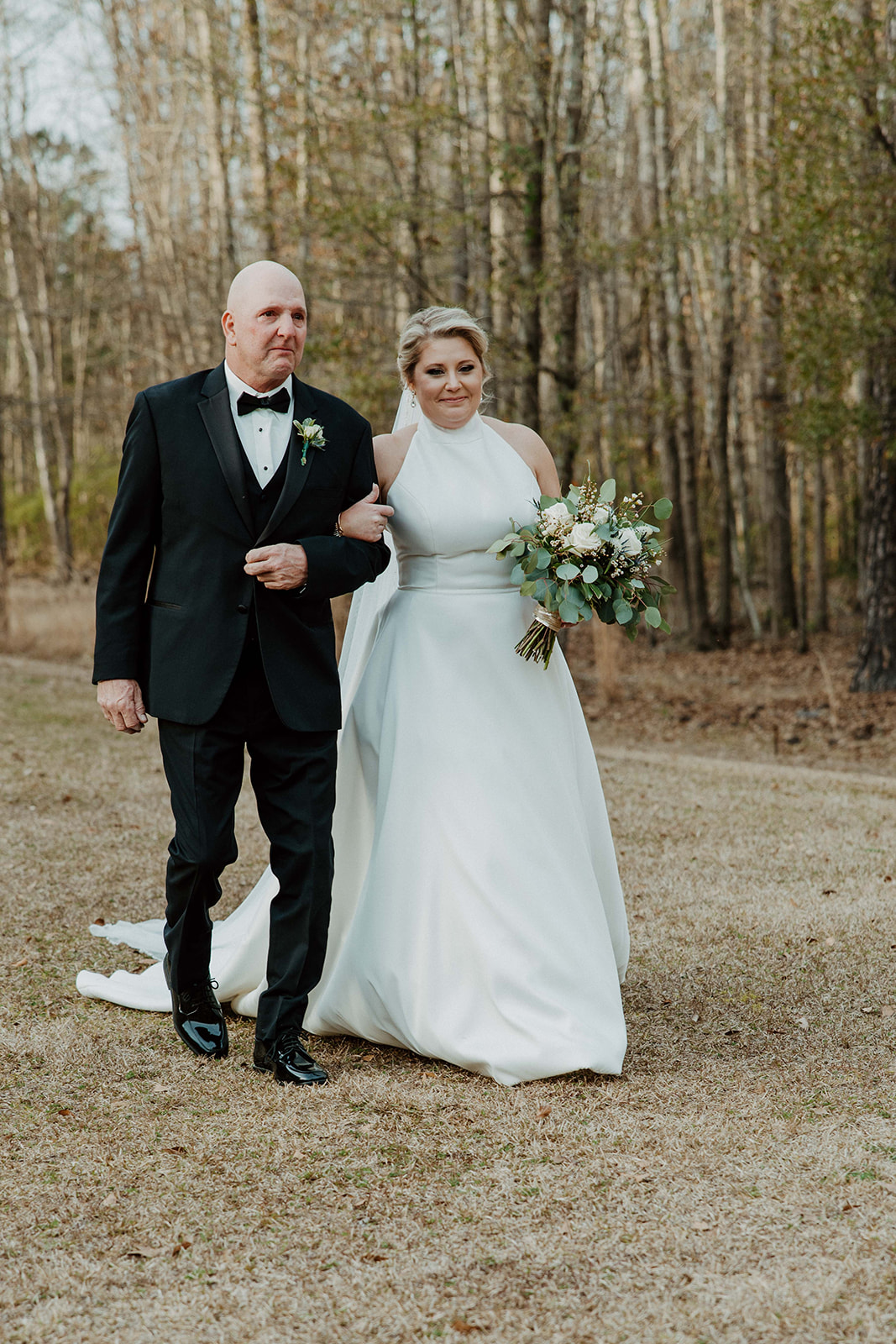 A bride and groom stand facing an officiant during their outdoor wedding ceremony, with a building visible in the background. Flowers are in the foreground at  The Venues at Ogeechee Tech
