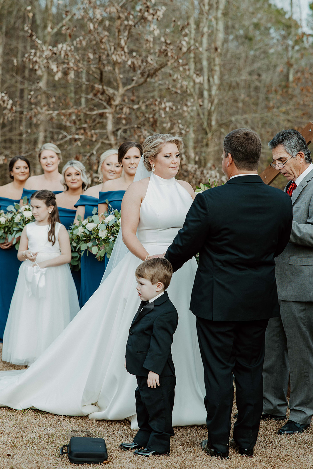 A bride and groom stand facing an officiant during their outdoor wedding ceremony, with a building visible in the background. Flowers are in the foreground at  The Venues at Ogeechee Tech
