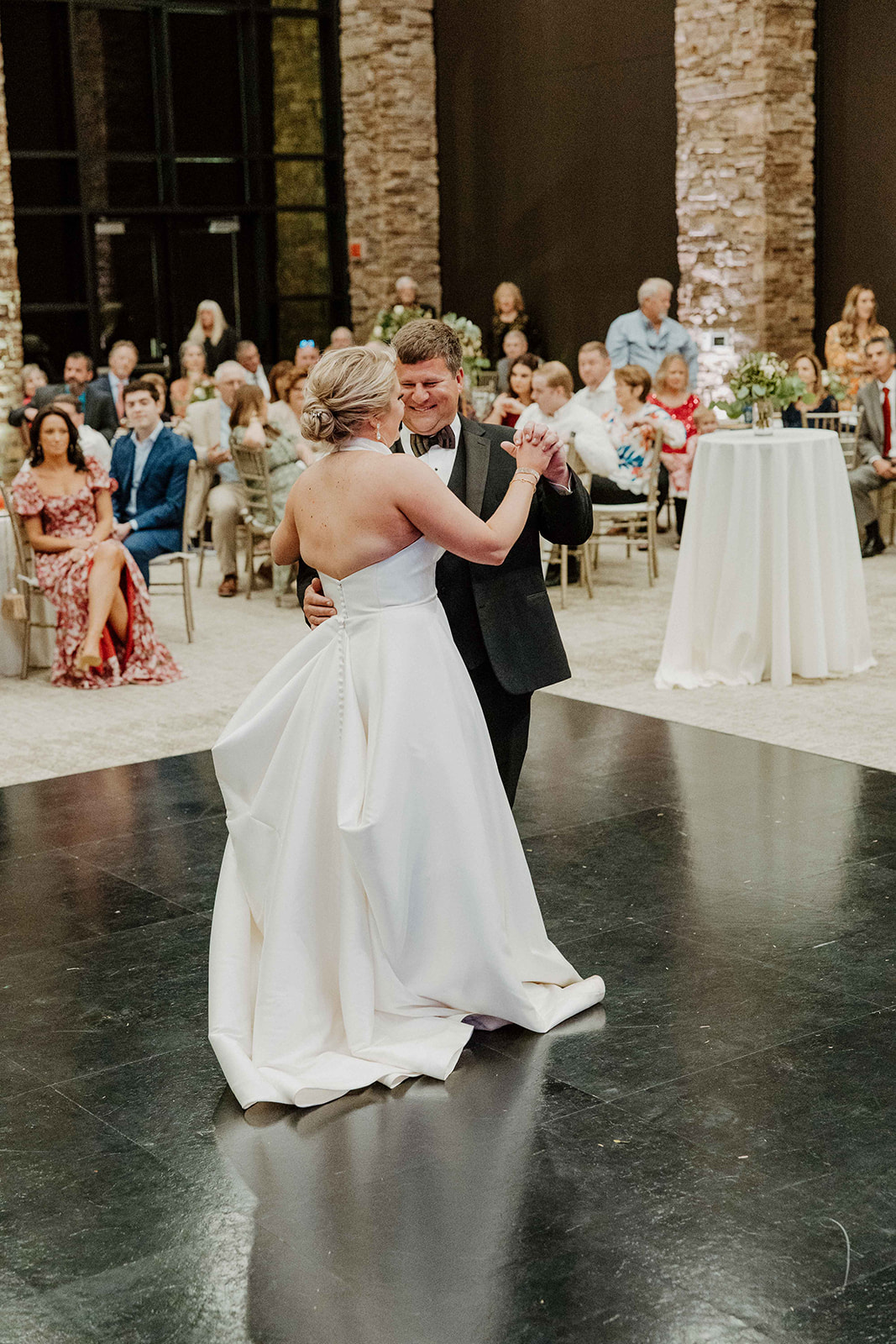 A couple dances on a black floor in a large, well-lit room with stone columns and seated guests watching them in the background. The woman is wearing a white gown, and the man is in a suit.