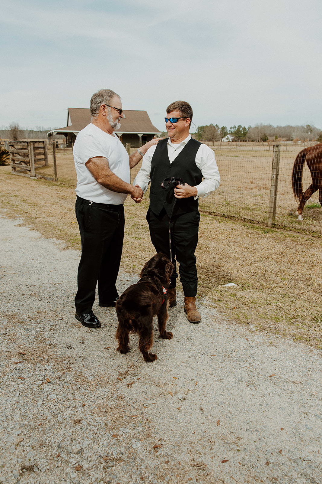 A woman helps a man adjust his tie outdoors. The man is wearing a suit and sunglasses. A dog on a leash sits in the foreground. Trees and a fence are in the background.