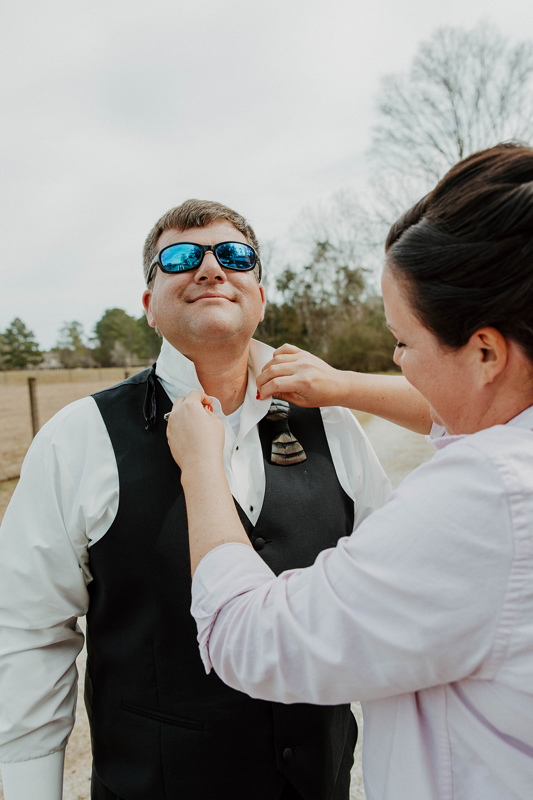 A woman helps a man adjust his tie outdoors. The man is wearing a suit and sunglasses. A dog on a leash sits in the foreground. Trees and a fence are in the background.