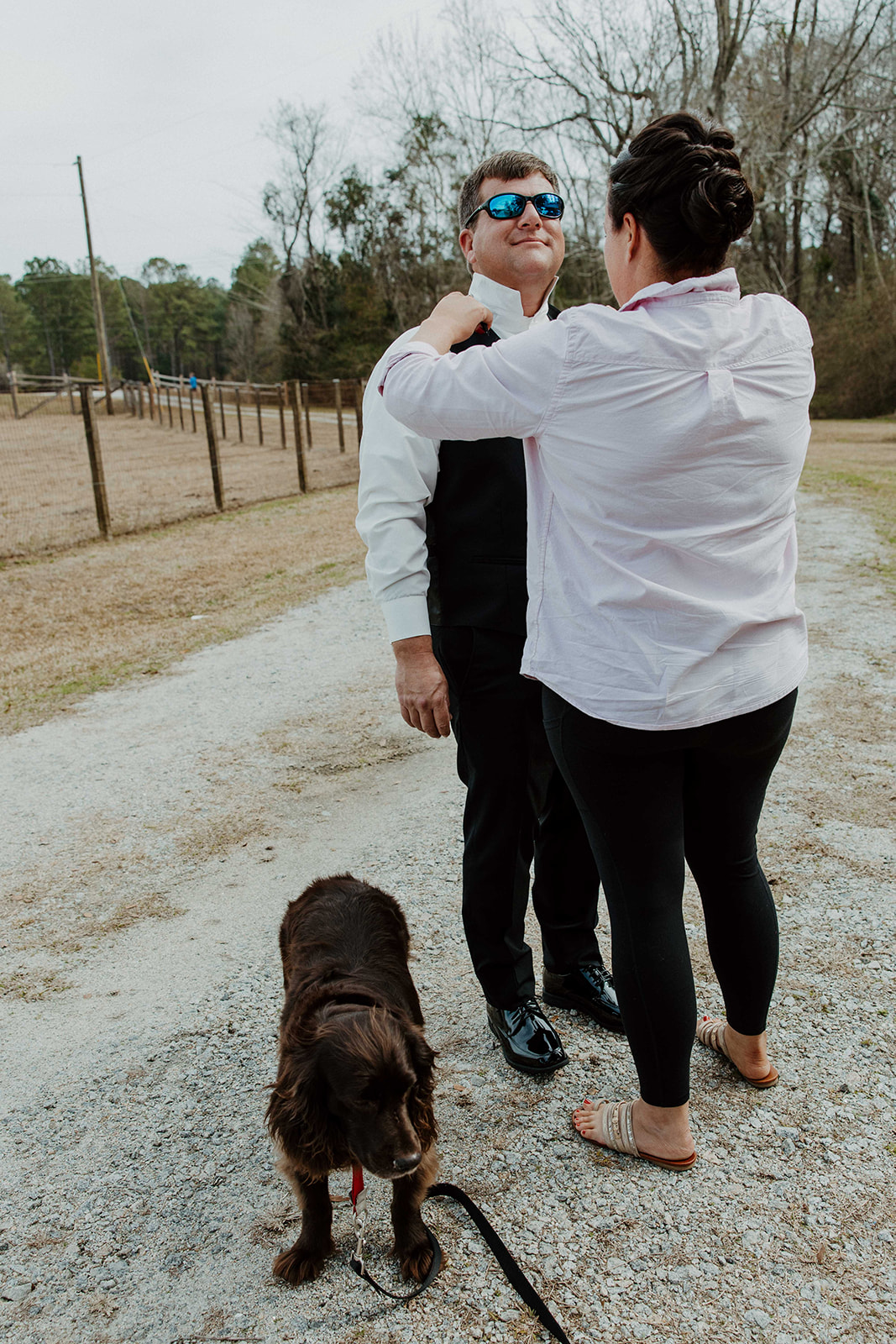 A woman helps a man adjust his tie outdoors. The man is wearing a suit and sunglasses. A dog on a leash sits in the foreground. Trees and a fence are in the background.