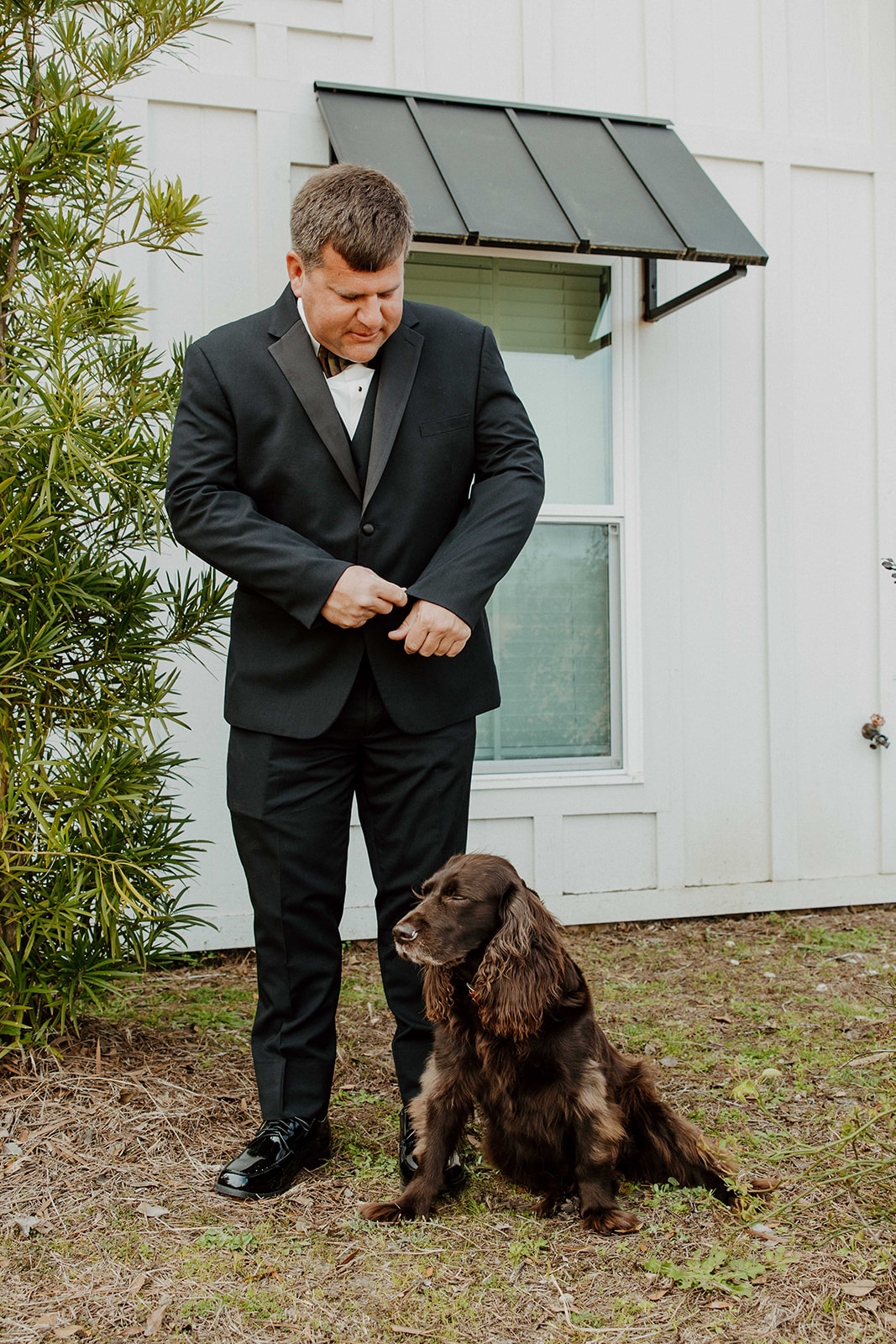 A man dressed in a black suit stands outside a white building, buttoning his jacket. A black and brown dog sits at his feet at The Venues at Ogeechee Tech
