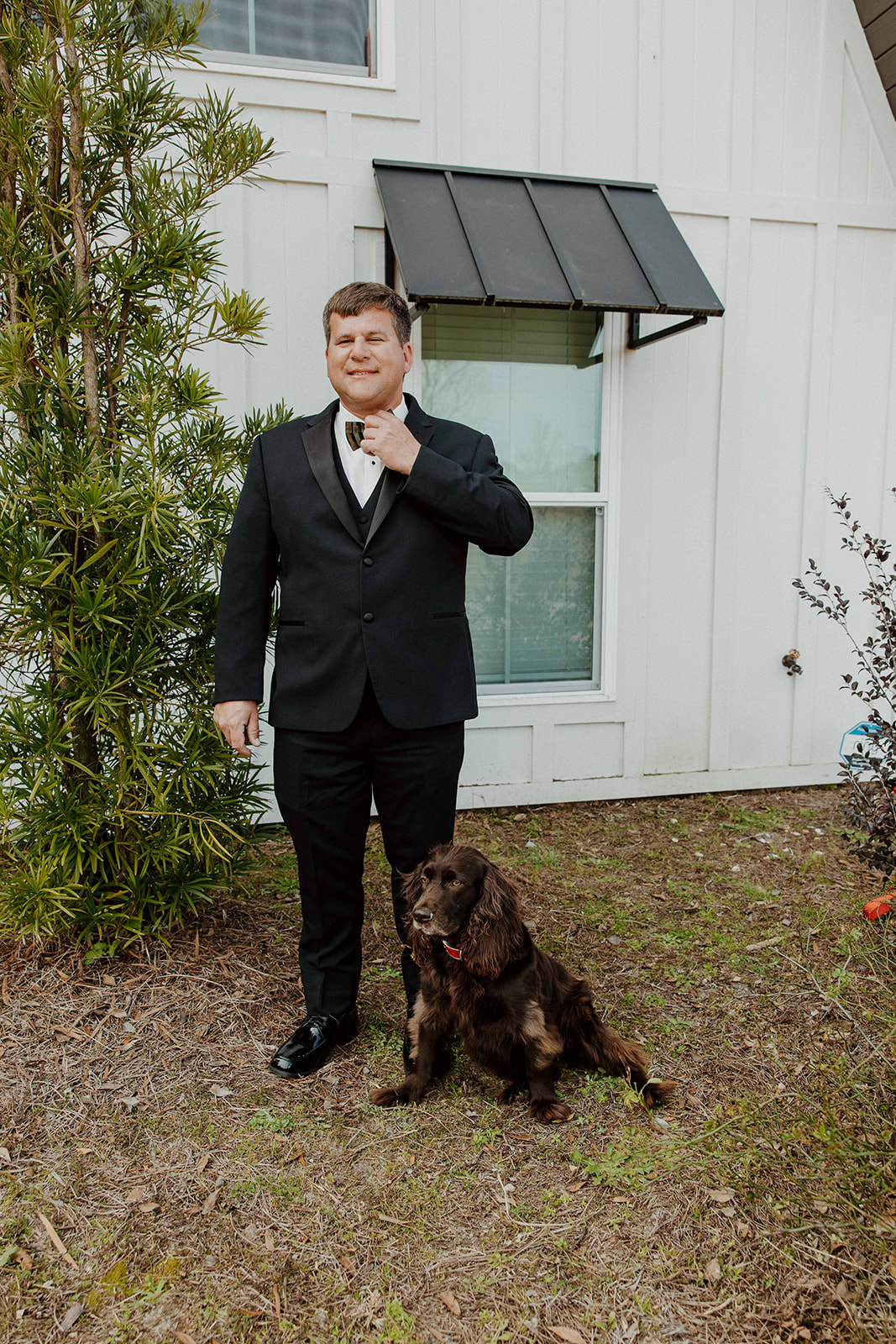 A man dressed in a black suit stands outside a white building, buttoning his jacket. A black and brown dog sits at his feet at The Venues at Ogeechee Tech
