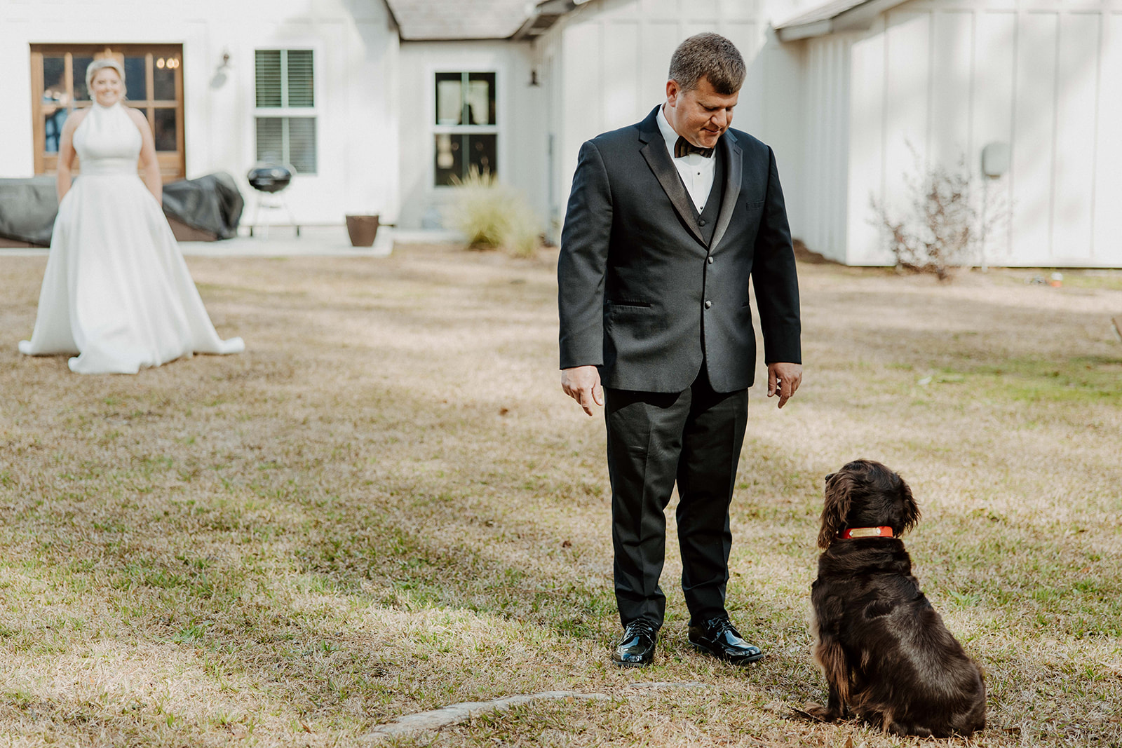 A bride in a white dress stands outside a white house, watching a man in a suit and bow tie walking a brown dog on a lawn during their first look at The Venues at Ogeechee Tech
