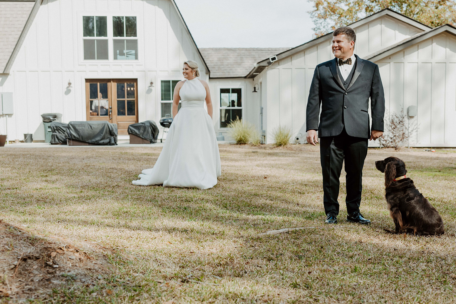 A bride in a white dress stands outside a white house, watching a man in a suit and bow tie walking a brown dog on a lawn during their first look at The Venues at Ogeechee Tech